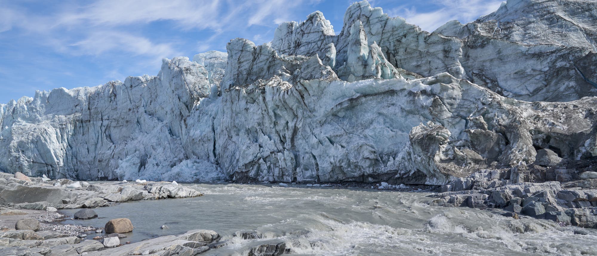 Gräuliches Schmelzwasser fließt über graues Gestein vor einer schmutzigweißen Eiswand in Grönland. Wenigstens ist der Himmel freundlich blau-weiß.