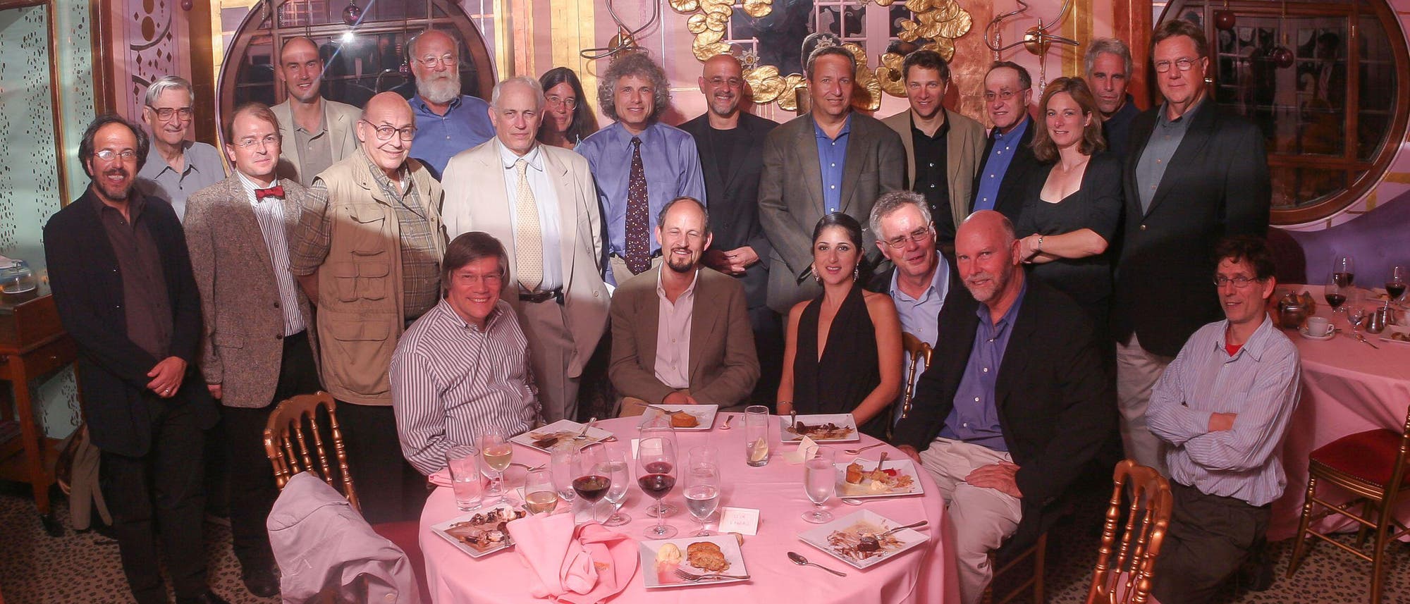 Eine Gruppe von Menschen posiert in einem eleganten Raum mit kunstvoller Wanddekoration. Sie stehen und sitzen um einen runden Tisch, der mit rosa Tischdecke und Geschirr gedeckt ist. Auf dem Tisch befinden sich Teller mit Speisen und Gl&auml;ser mit Getr&auml;nken. Die Personen l&auml;cheln und blicken in die Kamera, was auf eine gesellige Veranstaltung hindeutet. Im Hintergrund sind dekorative Elemente und runde Fenster zu sehen.