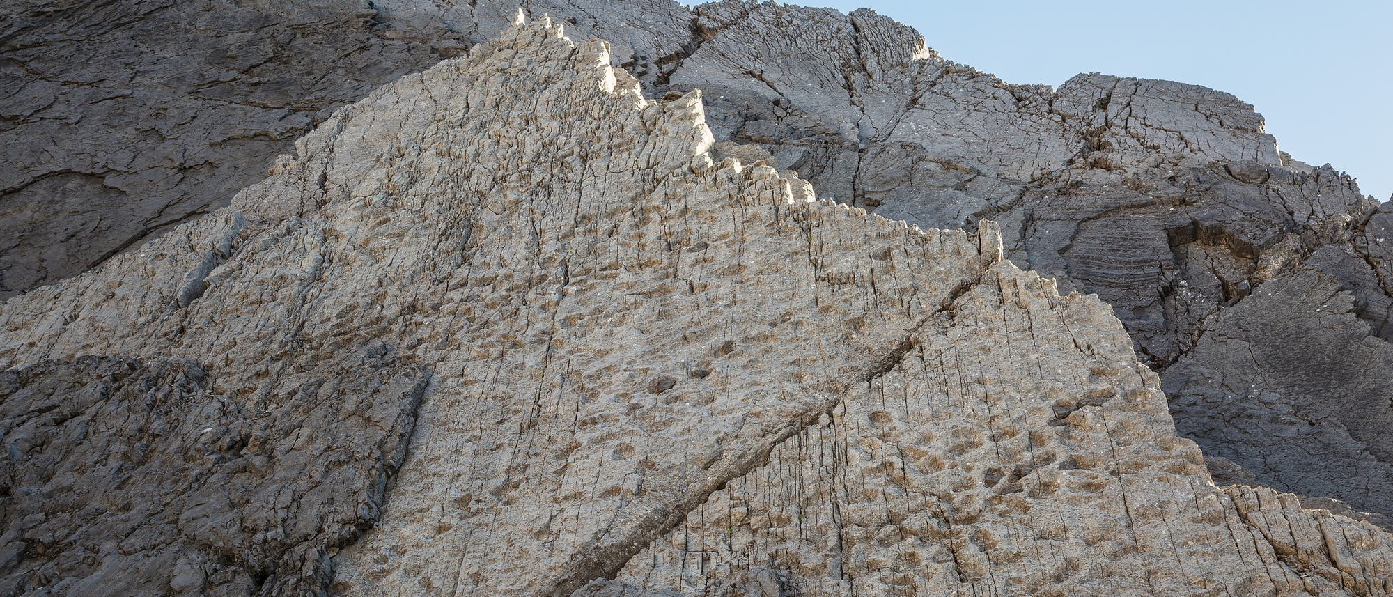 Felsformation mit markanten, schr&auml;g verlaufenden Schichten und Rissen. Die Oberfl&auml;che ist &uuml;bers&auml;t mit Abdr&uuml;cken und zeigt eine Mischung aus hellen und dunklen Gesteinsfarben, die auf geologische Prozesse hinweisen. Der Himmel ist klar und blau im Hintergrund.