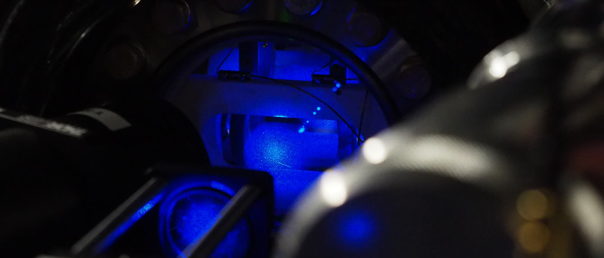 Eine Nahaufnahme eines wissenschaftlichen Ger&auml;ts, das in bl&auml;ulichem Licht leuchtet. Im Zentrum des Bilds befindet sich ein kreisf&ouml;rmiges Element, umgeben von metallischen Strukturen und Kabeln. Die Szene ist dunkel, mit einem Fokus auf die leuchtenden Teile des Ger&auml;ts, die m&ouml;glicherweise f&uuml;r Experimente oder Messungen verwendet werden.