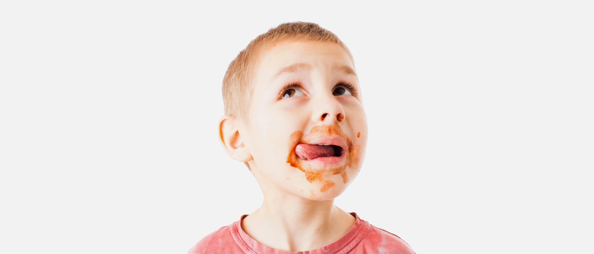 Ein kleiner Junge mit kurzen Haaren leckt sich die Lippen, während er nachdenklich nach oben schaut. Sein Mund ist mit Schokoladenflecken verschmiert. Er trägt ein rotes T-Shirt. Der Hintergrund ist einfarbig hellgrau.