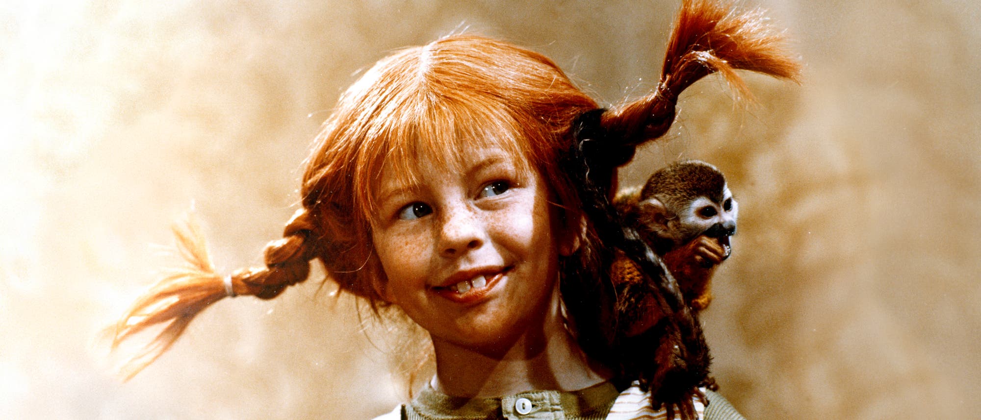 Ein Kind mit roten Zöpfen lächelt und schaut zur Seite. Auf der Schulter des Kindes sitzt ein kleiner Affe. Der Hintergrund ist unscharf und in warmen Tönen gehalten.