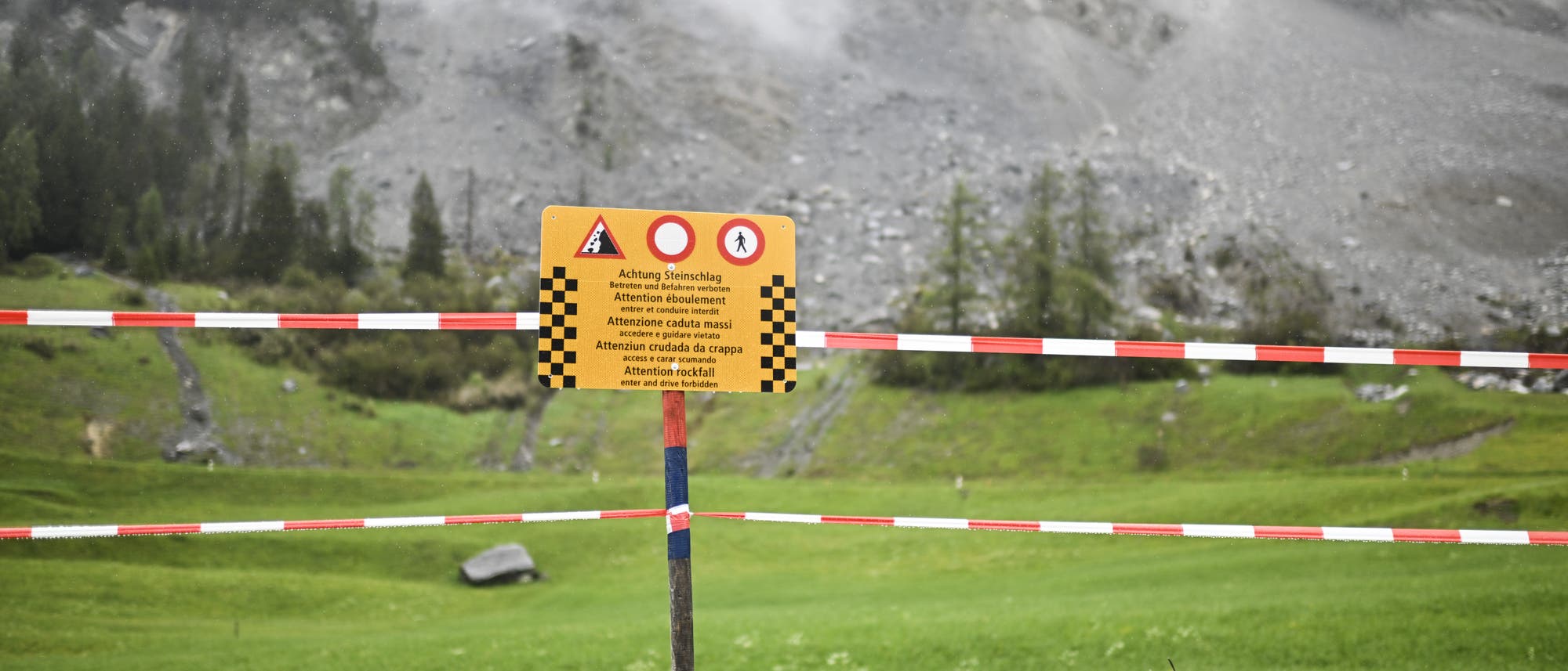 Ein gelbes Warnschild warnt davor, das dahinter liegende Gebiet zu betreten, weil ein Bergsturz droht. Verschwommen sieht man im Hintergrund den grauen Fels und eine Felsschutthalde. Beides ist umgeben von grünen Wiesen und Hängen.