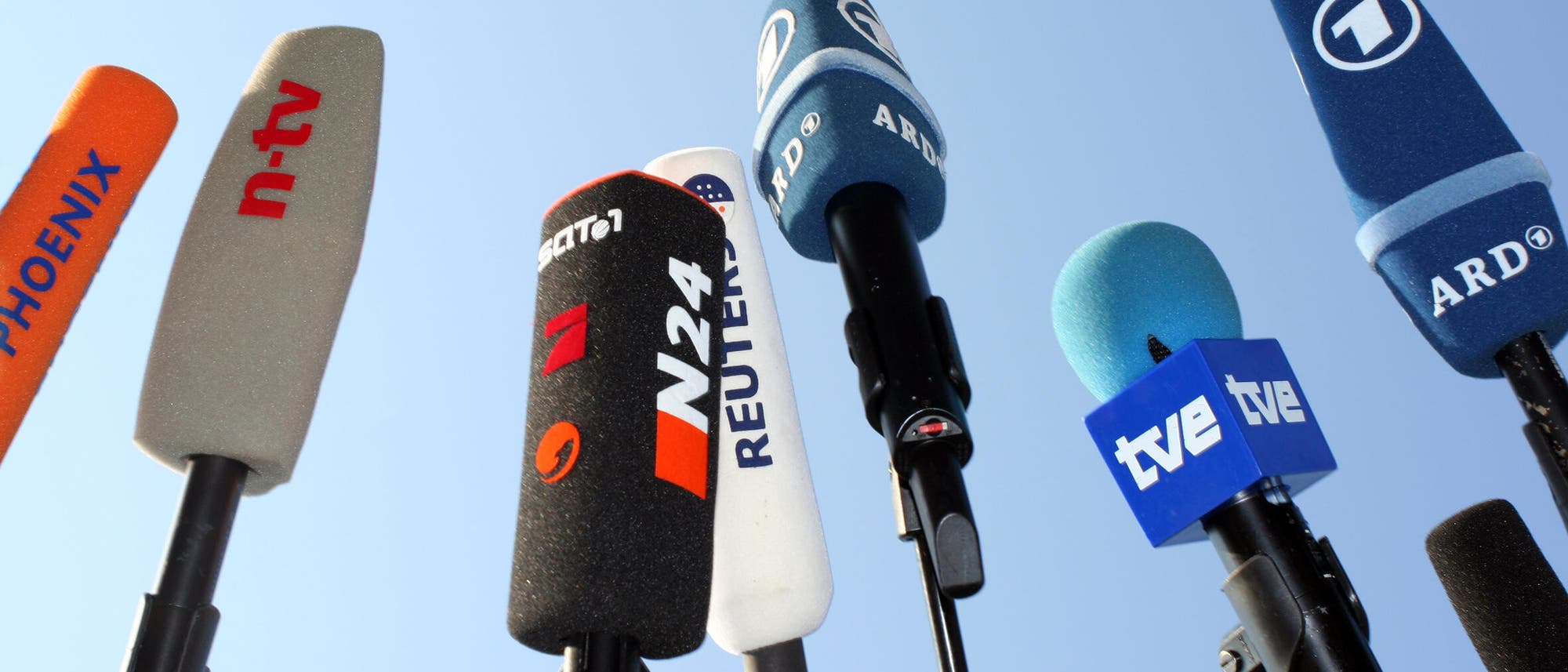 Mehrere Mikrofone verschiedener Nachrichtensender sind in die Luft gerichtet, darunter Phoenix, n-tv, N24, Reuters, ARD und tve. Die Mikrofone sind mit ihren jeweiligen Logos gekennzeichnet und stehen vor einem klaren blauen Himmel, was auf eine Pressekonferenz oder ein Medienereignis hindeutet. Mehrere Mikrofone verschiedener Nachrichtensender sind in die Luft gerichtet, darunter Phoenix, n-tv, N24, Reuters, ARD und tve. Die Mikrofone sind mit ihren jeweiligen Logos gekennzeichnet und stehen vor einem klaren blauen Himmel, was auf eine Pressekonferenz oder ein Medienereignis hindeutet.