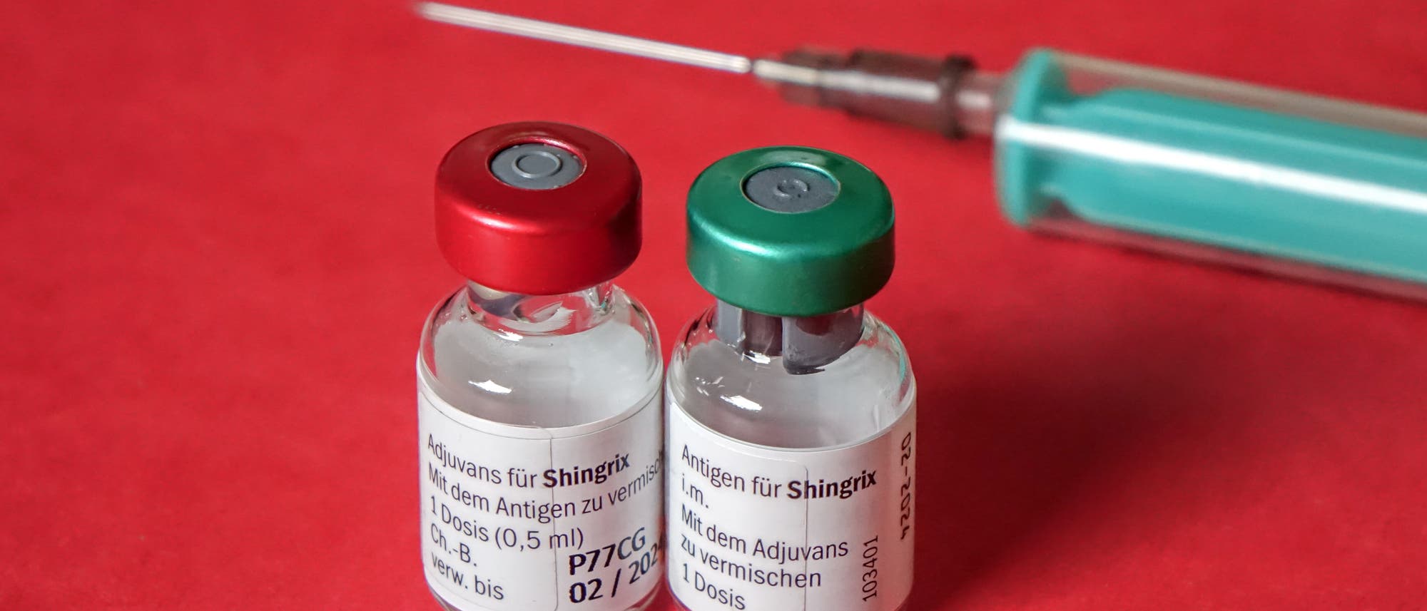 Zwei Impfstofffläschchen mit der Aufschrift „Adjuvans für Shingrix“ und „Antigen für Shingrix“ stehen nebeneinander auf einer roten Oberfläche. Die Fläschchen haben einen roten beziehungsweise grünen Verschluss. Im Hintergrund ist eine Spritze mit einer langen Nadel zu sehen.