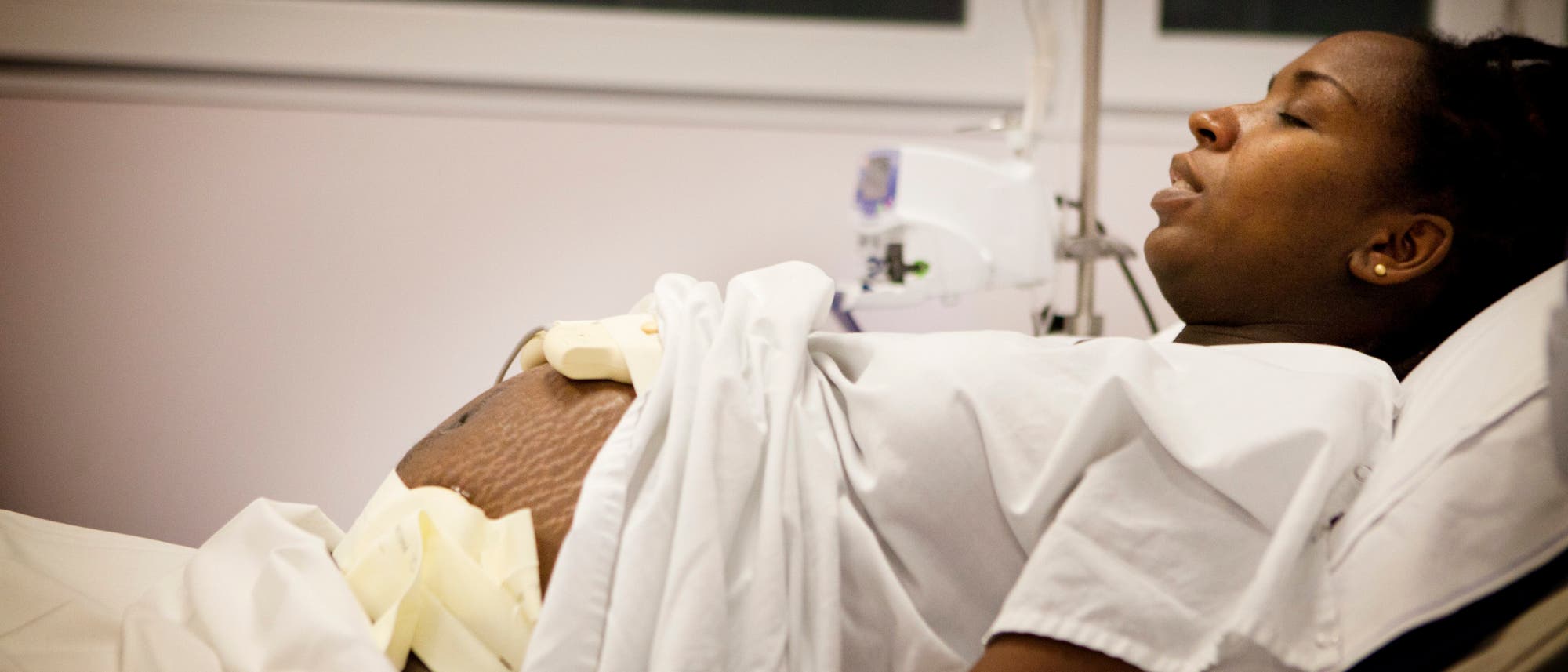 Eine schwangere Frau liegt in einem Krankenhausbett, teilweise zugedeckt mit einem weißen Laken. Sie trägt ein Krankenhaushemd und scheint sich in einer Entbindungsstation zu befinden. Neben ihr ist medizinisches Equipment sichtbar. Die Frau wirkt konzentriert und ruhig.