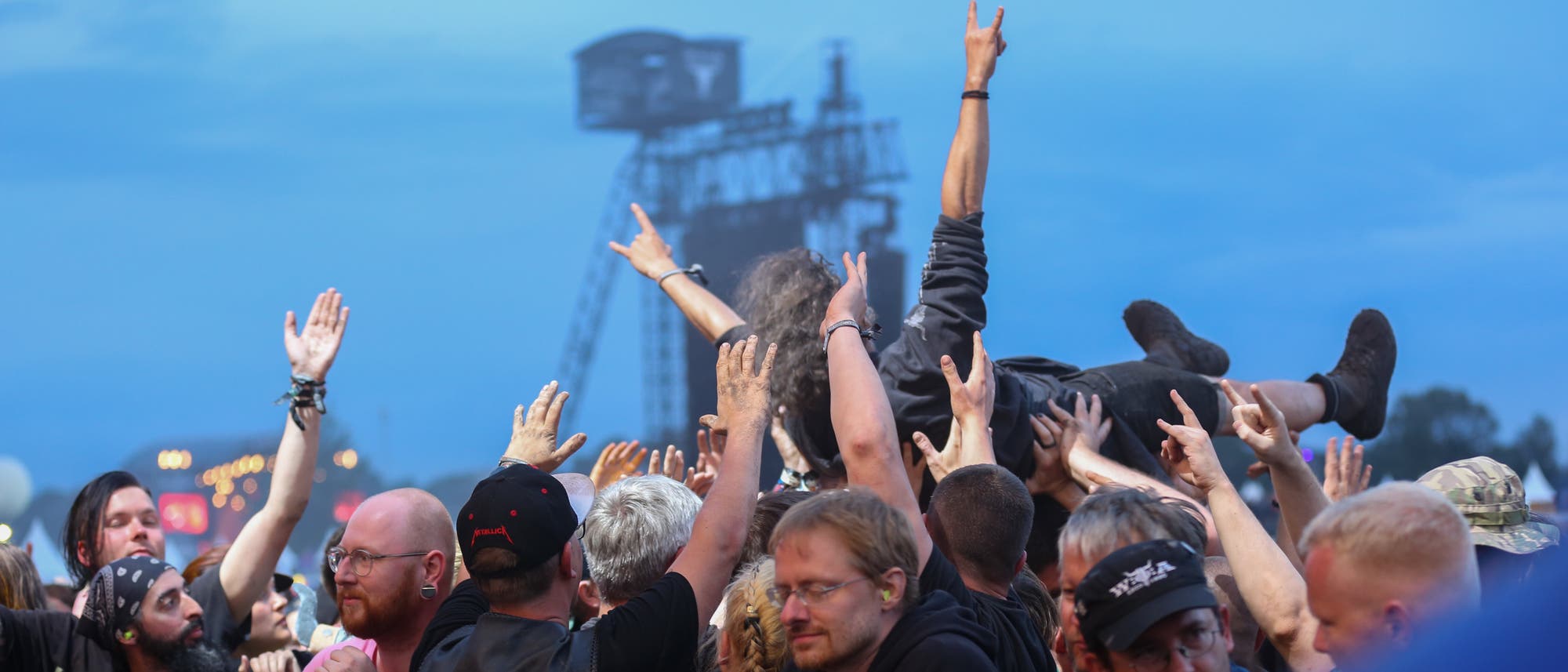 Eine Menschenmenge bei einem Konzert auf dem Wacken Open Air hebt eine Person zum Crowdsurfing über ihre Köpfe. Die Szene findet vor einer großen Bühne statt. Viele der Anwesenden tragen Band-T-Shirts und zeigen Handgesten, die oft bei Rockkonzerten verwendet werden. Eine Menschenmenge bei einem Konzert auf dem Wacken Open Air hebt eine Person zum Crowdsurfing über ihre Köpfe. Die Szene findet vor einer großen Bühne statt. Viele der Anwesenden tragen Band-T-Shirts und zeigen Handgesten, die oft bei Rockkonzerten verwendet werden.