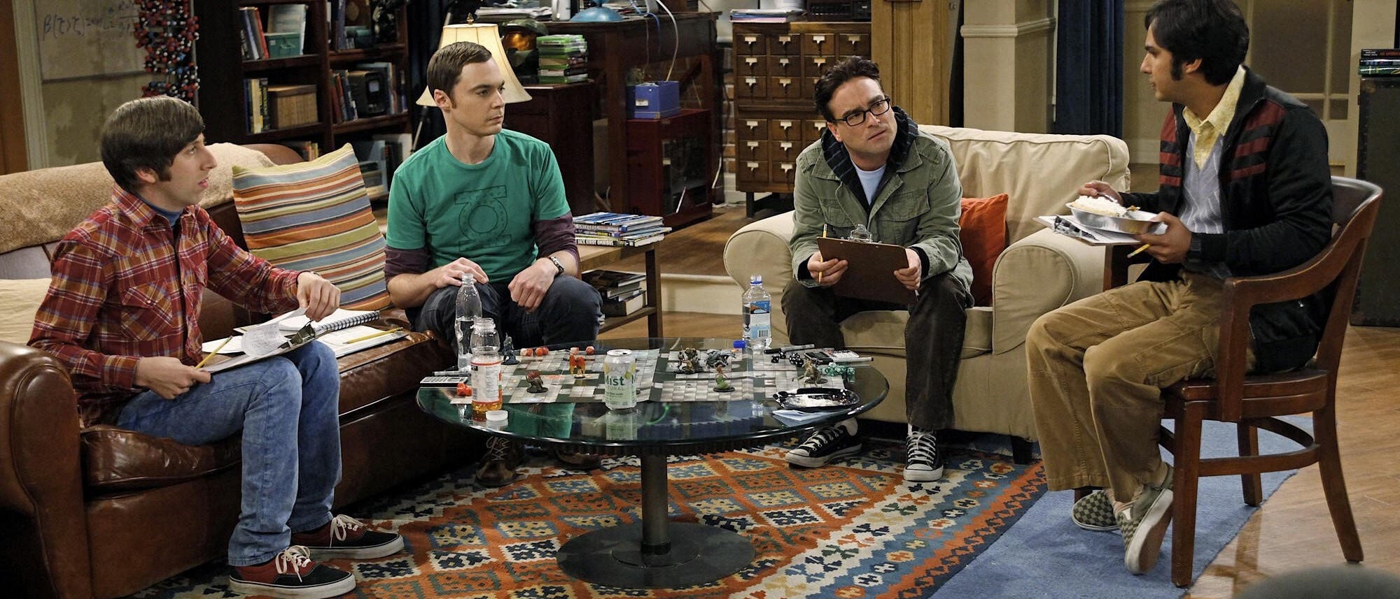 Eine Gruppe von vier Personen sitzt in einem Wohnzimmer um einen runden Glastisch, der mit verschiedenen Spielmaterialien bedeckt ist. Im Hintergrund sind B&uuml;cherregale, ein Teleskop und wissenschaftliche Poster zu sehen. Die Personen scheinen in ein Gespr&auml;ch vertieft zu sein, w&auml;hrend sie Notizbl&ouml;cke und Stifte halten. Der Raum ist gem&uuml;tlich eingerichtet mit einem gemusterten Teppich und einem Sofa.