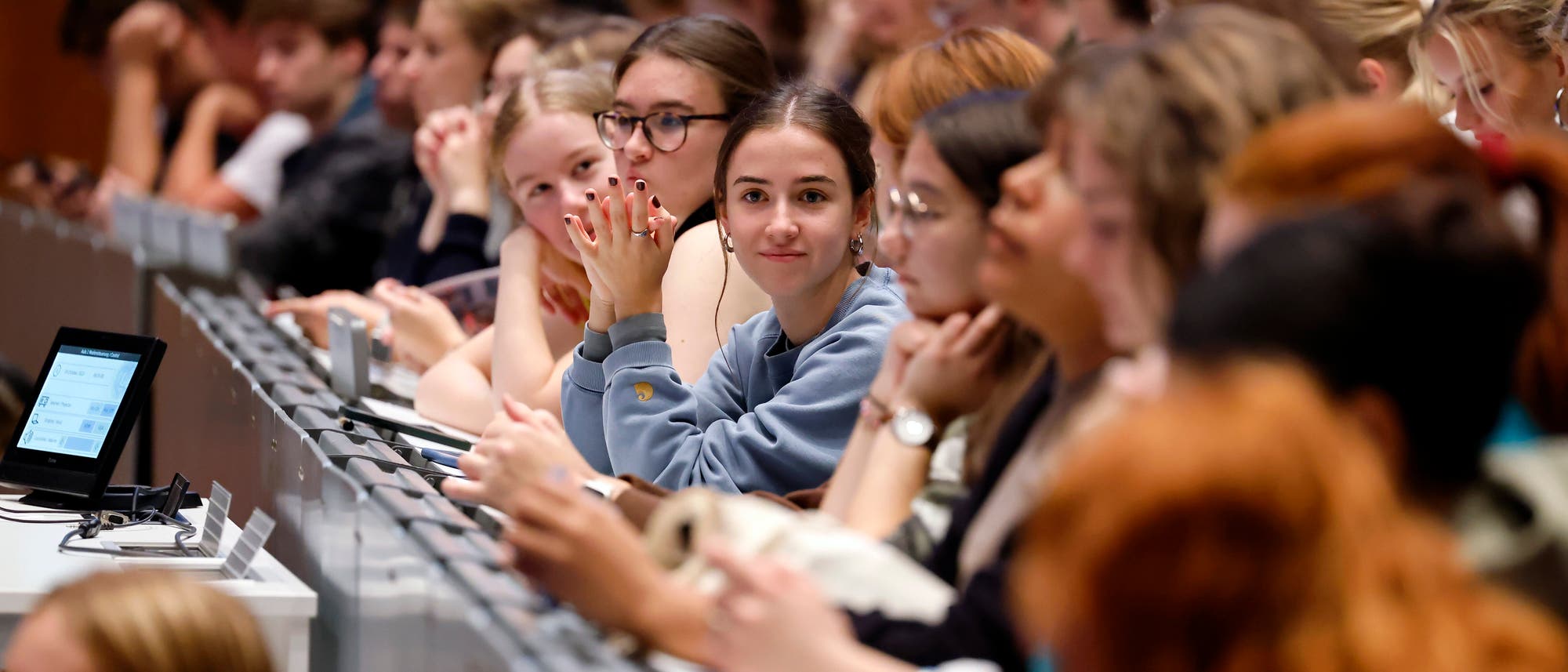 Eine Gruppe von Studierenden sitzt in einem Hörsaal. Eine junge Frau in der Mitte mit einem blauen Pullover schaut direkt in die Kamera. Vor ihr befindet sich ein Tablet mit Notizen. Die Atmosphäre ist aufgekratzt und lernorientiert.