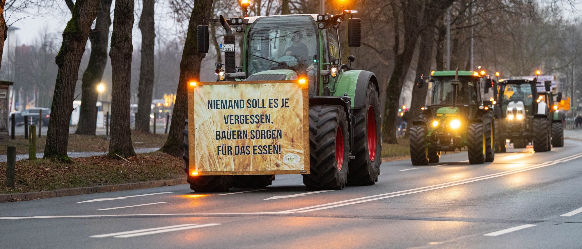 Mehrere Traktoren fahren hintereinander auf der Straße. Der vorderste hat ein Schild an der Stoßstange, auf dem steht: Niemand soll es je vergessen, Bauern sorgen für das Essen.