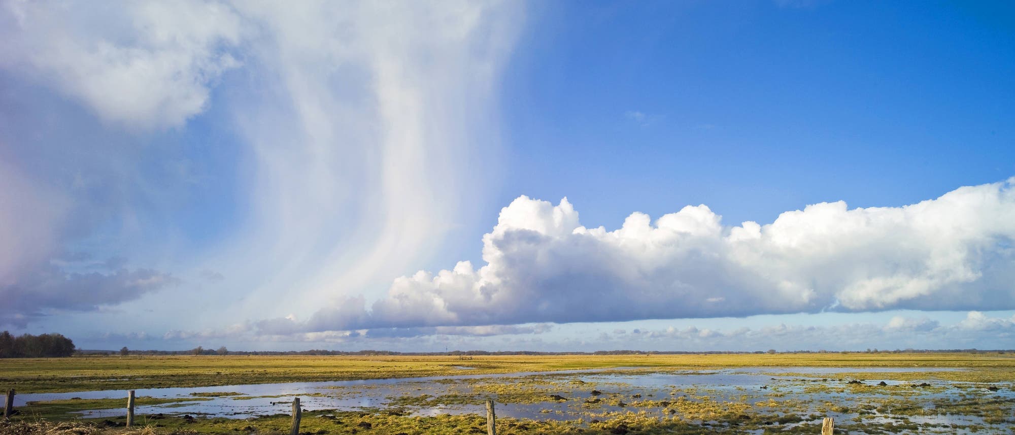Eine weitl&auml;ufige Landschaft mit einem klaren blauen Himmel und dramatischen Wolkenformationen. Im Vordergrund sind feuchte Wiesen und ein Wassergraben zu sehen, die von einem niedrigen Zaun begrenzt werden. Die Wolken werfen Schatten auf die Wiesen, was ein Spiel von Licht und Schatten erzeugt.