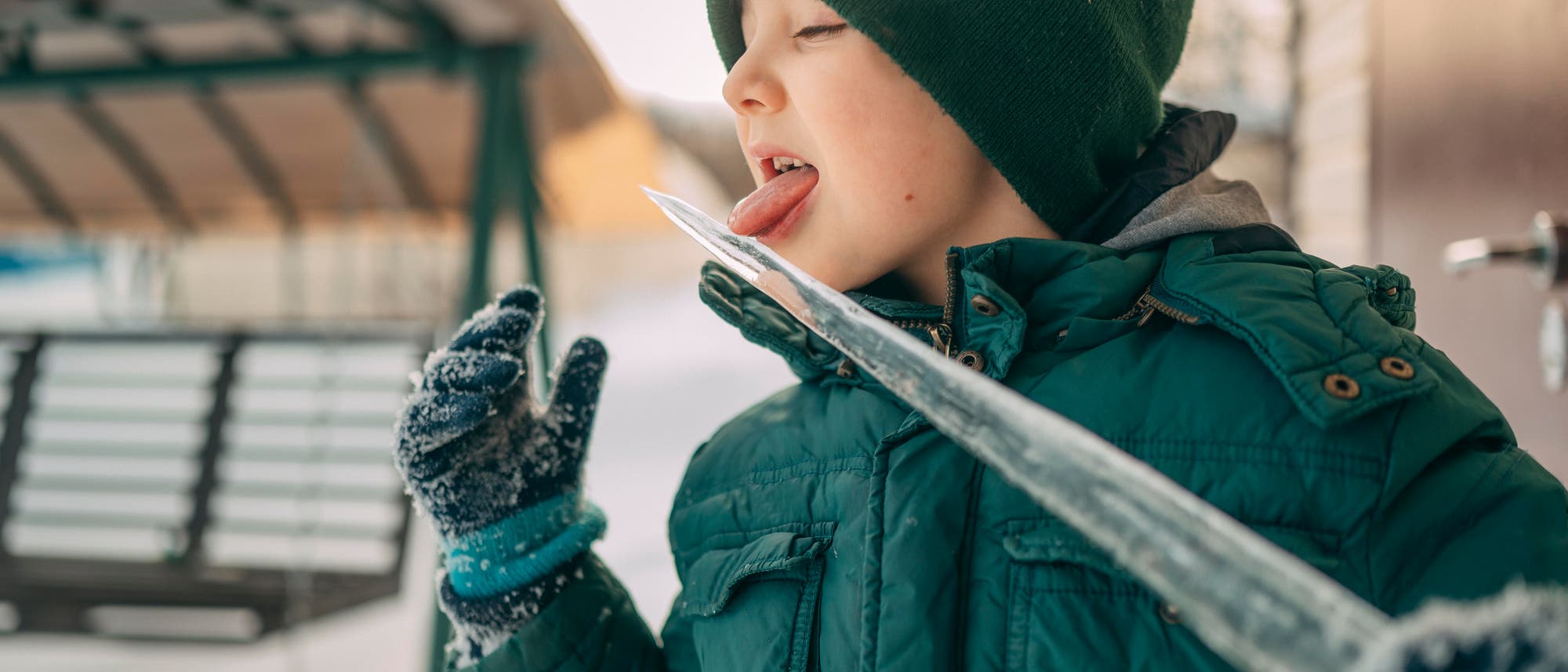 Ein Kind in Winterkleidung, einschlie&szlig;lich einer gr&uuml;nen M&uuml;tze und einer dicken Jacke, steht drau&szlig;en im Schnee. Es streckt die Zunge aus, um an einem gro&szlig;en Eiszapfen zu lecken. Im Hintergrund ist eine schneebedeckte Schaukel zu sehen. Die Szene vermittelt eine spielerische Winteratmosph&auml;re.