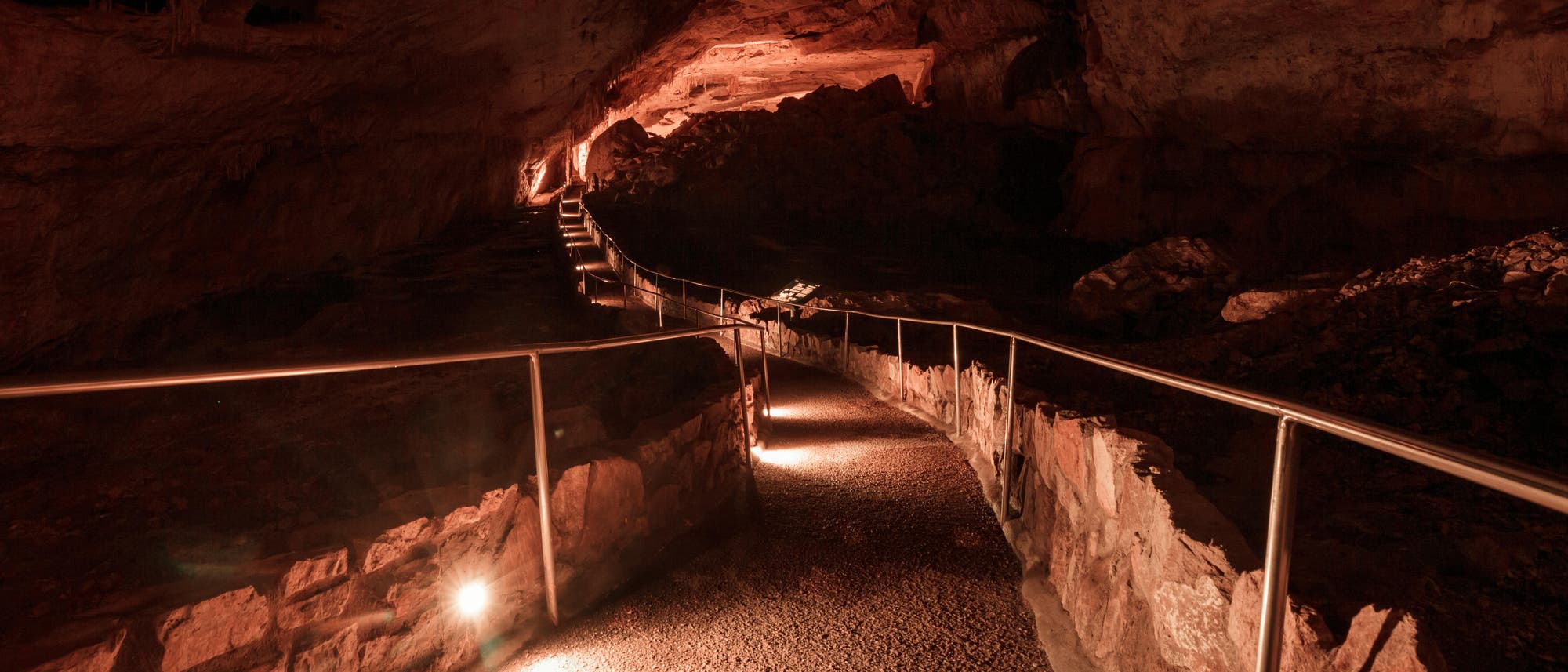 Ein beleuchteter Weg führt durch die Carlsbad Caverns in New Mexico. Der Weg ist grobkörnig und grau und von gemauerten Steinen eingefasst, ein Handlauf aus Stahl mit stählernen Pfosten führt an ihm entlang. Die Höhlendecke wirkt im Licht rötlich. Insgesamt ist es düster.