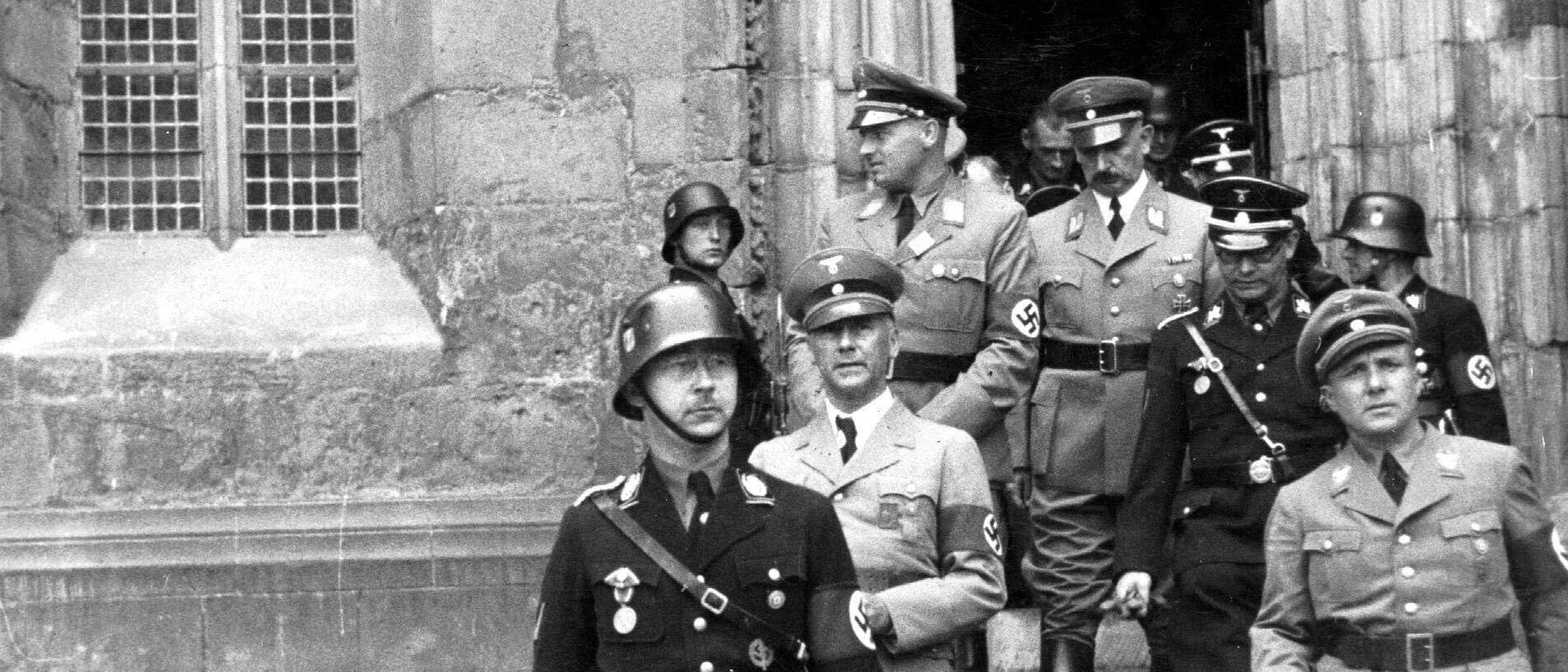 Eine Gruppe von Männern in militärischen Uniformen verlässt ein historisches Gebäude mit gotischen Fenstern und einem verzierten Eingang. Die Männer tragen Helme und Abzeichen, während sie die Steinstufen hinabsteigen. Die Szene vermittelt einen formellen und ernsten Eindruck. Eine Gruppe von Männern in militärischen Uniformen verlässt ein historisches Gebäude mit gotischen Fenstern und einem verzierten Eingang. Die Männer tragen Helme und Abzeichen, während sie die Steinstufen hinabsteigen. Die Szene vermittelt einen formellen und ernsten Eindruck.