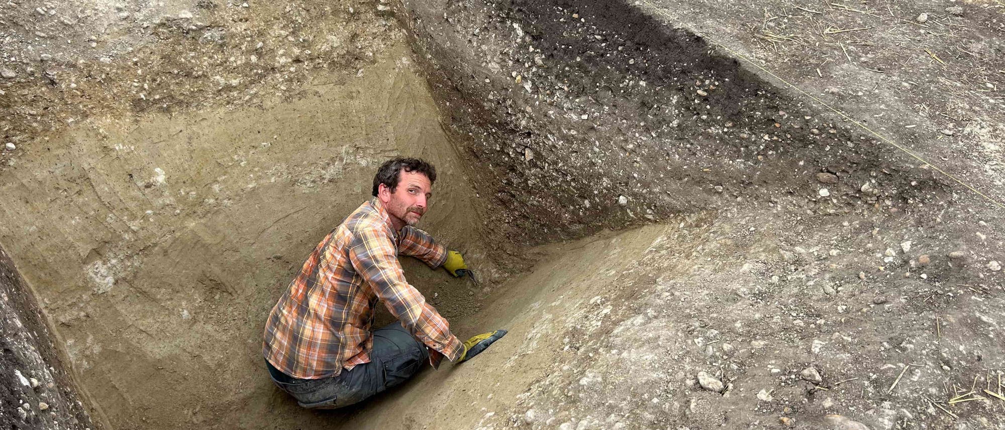 Eine Person in einem karierten Hemd und Handschuhen gr&auml;bt in einer arch&auml;ologischen Ausgrabungsst&auml;tte. Sie befindet sich in einer tiefen, spitzf&ouml;rmigen Grube und verwendet ein kleines Werkzeug, um den Boden zu bearbeiten. Die Umgebung zeigt mehrere Erdschichten und freigelegte Bereiche, die auf eine systematische Untersuchung hindeuten.