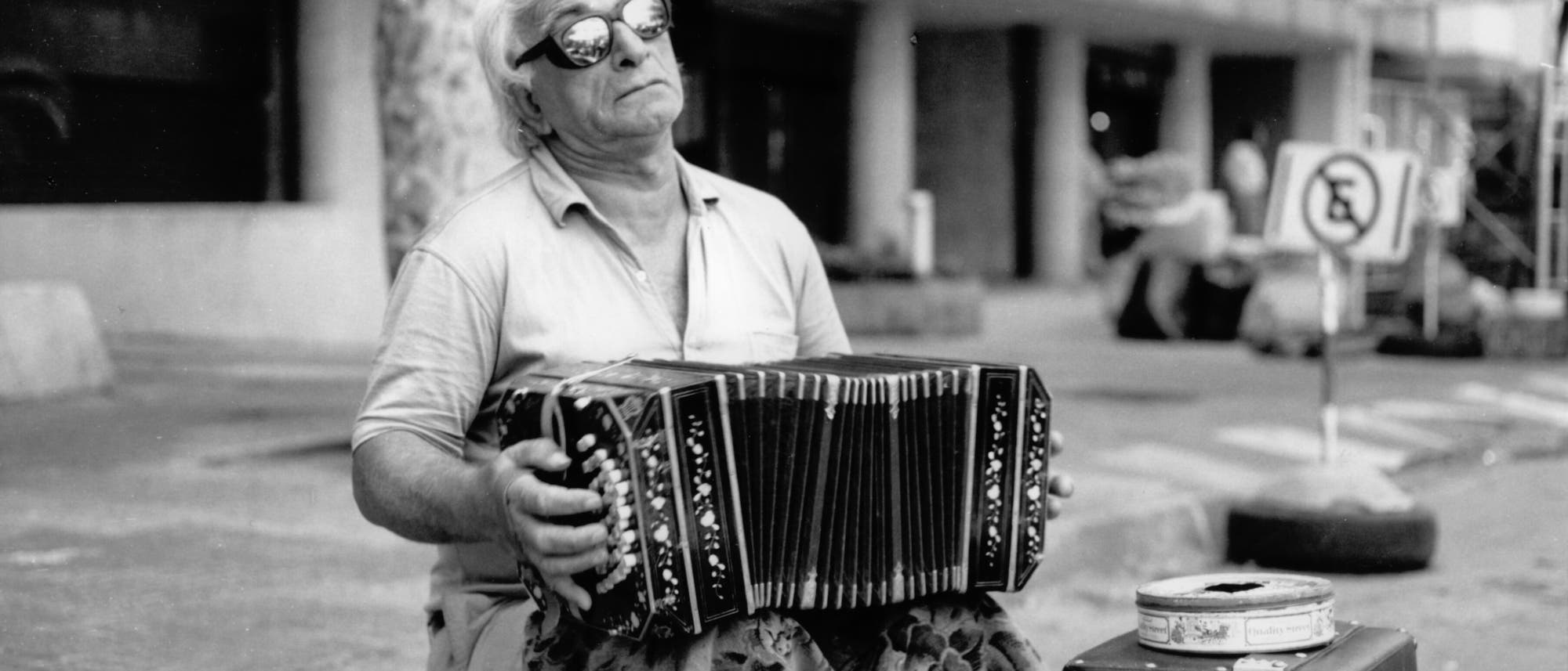 Ein älterer Mann mit Sonnenbrille sitzt auf einem Bürgersteig und spielt ein Bandoneon. Er trägt ein helles Hemd und hat graues Haar. Neben ihm steht ein offener Koffer mit einer Blechdose darauf. Im Hintergrund sind unscharfe Gebäude und ein Verkehrsschild zu sehen. Die Szene vermittelt eine Straßenmusikatmosphäre. Ein älterer Mann mit Sonnenbrille sitzt auf einem Bürgersteig und spielt ein Bandoneon. Er trägt ein helles Hemd und hat graues Haar. Neben ihm steht ein offener Koffer mit einer Blechdose darauf. Im Hintergrund sind unscharfe Gebäude und ein Verkehrsschild zu sehen. Die Szene vermittelt eine Straßenmusikatmosphäre.
