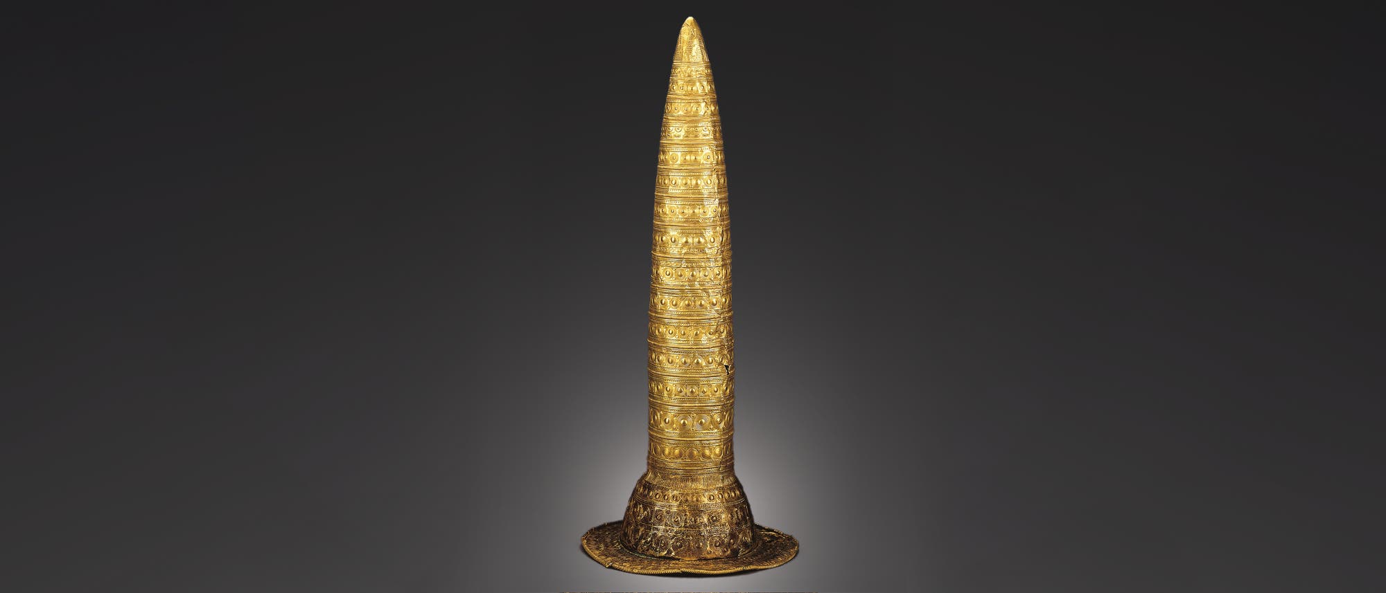 Ein konischer, goldener Hut mit detaillierten, kreisförmigen und linearen Mustern, die die Oberfläche bedecken. Der Hut hat eine breite Krempe und ist auf einem neutralen, dunklen Hintergrund abgebildet. Die Verzierungen sind symmetrisch und gleichmäßig verteilt, was auf eine kunstvolle Handwerkskunst hinweist.