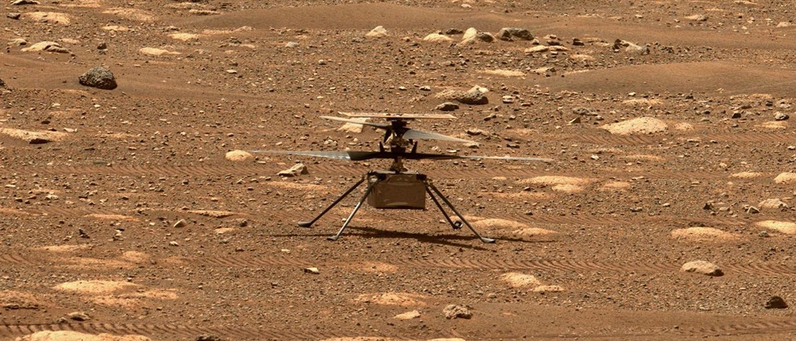Ein kleiner Helikopter steht auf der felsigen, r&ouml;tlichen Oberfl&auml;che des Mars. Der Boden ist mit kleinen Steinen und Spuren von Fahrzeugr&auml;dern bedeckt. Der Helikopter hat zwei Rotorbl&auml;tter und vier d&uuml;nne Beine. Die Szene zeigt die karge, staubige Landschaft des Mars.