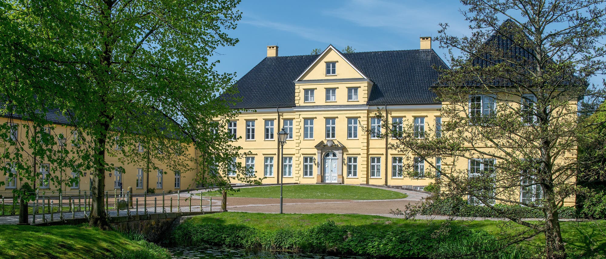 Ein großes, gelbes Herrenhaus mit schwarzen Dächern spiegelt sich in einem ruhigen Teich wider. Es handelt sich um das Landesarchiv Schleswig-Holstein. Das Gebäude ist von grünen Bäumen umgeben, die einen friedlichen Park umrahmen. Ein gepflasterter Weg führt zum Eingang des Hauses. Die Szene vermittelt eine ruhige, idyllische Atmosphäre. Ein großes, gelbes Herrenhaus mit schwarzen Dächern spiegelt sich in einem ruhigen Teich wider. Es handelt sich um das Landesarchiv Schleswig-Holstein. Das Gebäude ist von grünen Bäumen umgeben, die einen friedlichen Park umrahmen. Ein gepflasterter Weg führt zum Eingang des Hauses. Die Szene vermittelt eine ruhige, idyllische Atmosphäre.