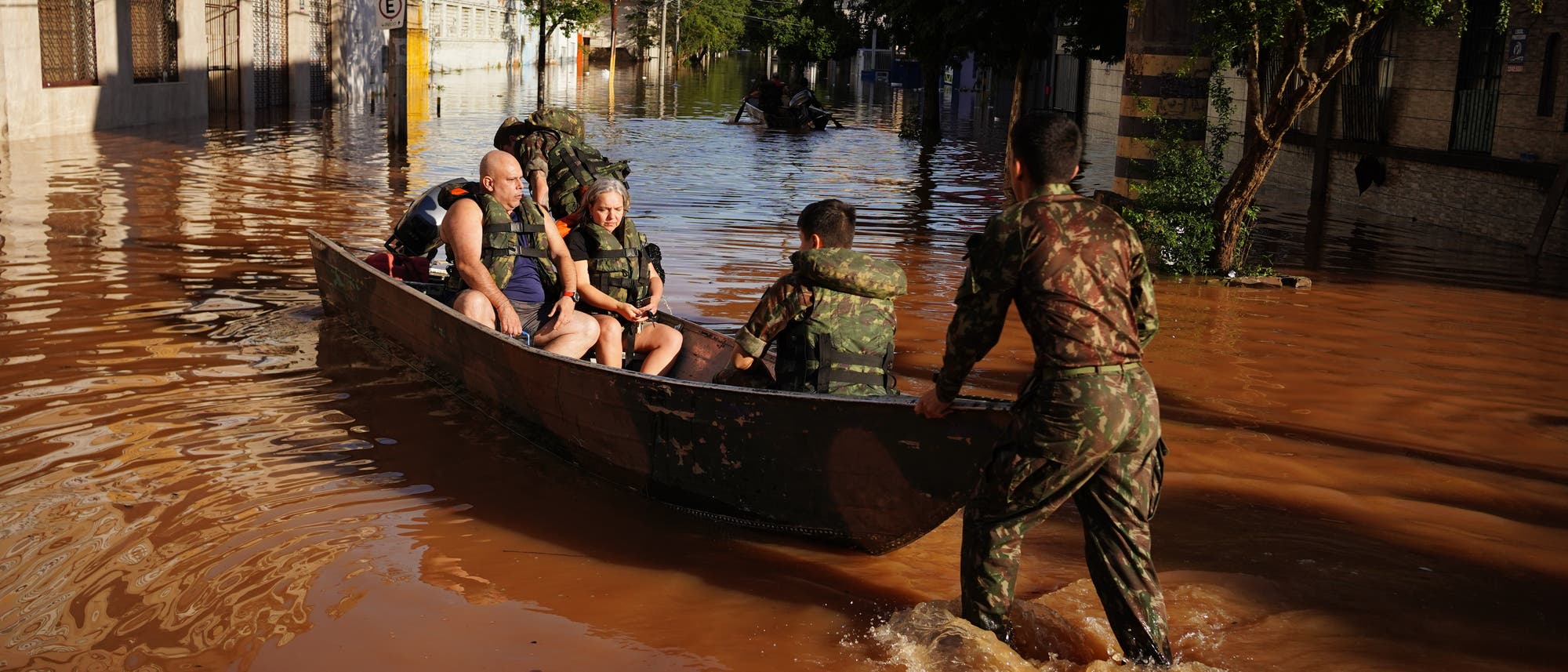 Menschen werden aufgrund der Überschwemmungen durch Mitarbeiter der brasilianischen Streitkräfte mit einem Boot evakuiert.  In den Straßen stehen braune Fluten, immerhin scheint gerade die Sonne.