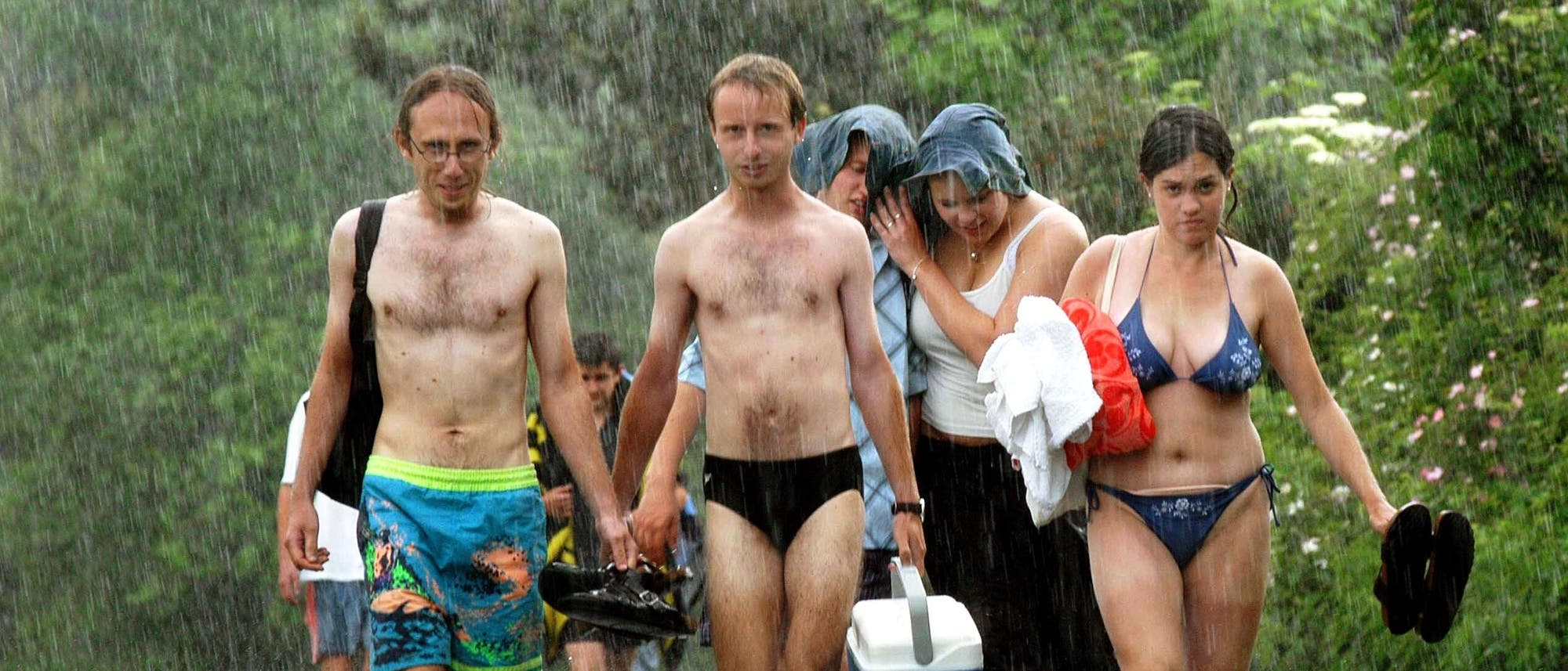 Eine Gruppe von fünf Menschen in Badebekleidung läuft im Regen einen Weg entlang. Zwei Männer tragen Badehosen, einer hält Schuhe in der Hand. Zwei Frauen schützen sich mit einem Handtuch über dem Kopf, während die dritte Frau ein Handtuch und Schuhe trägt. Im Hintergrund ist grüne Vegetation zu sehen. Die Stimmung wirkt unbeschwert trotz des Sommerregens. Eine Gruppe von fünf Menschen in Badebekleidung läuft im Regen einen Weg entlang. Zwei Männer tragen Badehosen, einer hält Schuhe in der Hand. Zwei Frauen schützen sich mit einem Handtuch über dem Kopf, während die dritte Frau ein Handtuch und Schuhe trägt. Im Hintergrund ist grüne Vegetation zu sehen. Die Stimmung wirkt unbeschwert trotz des Sommerregens.