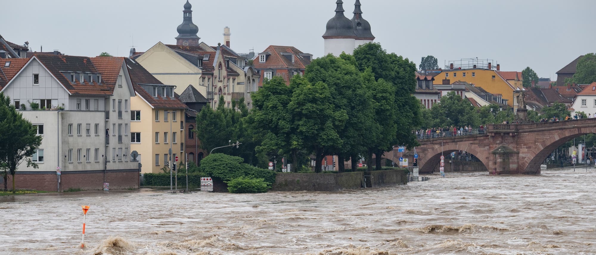 Die braunen Fluten des Neckars schießen durch Heidelberg und überfluten Teile der Altstadt. Der Blick geht von Osten in Richtung alte Brücke, auf der zahlreiche menschen stehen. Links ist die Altstadt mit verschiedenen Gebäuden zu sehen, darunter auch der Turm der Heiliggeistkirche