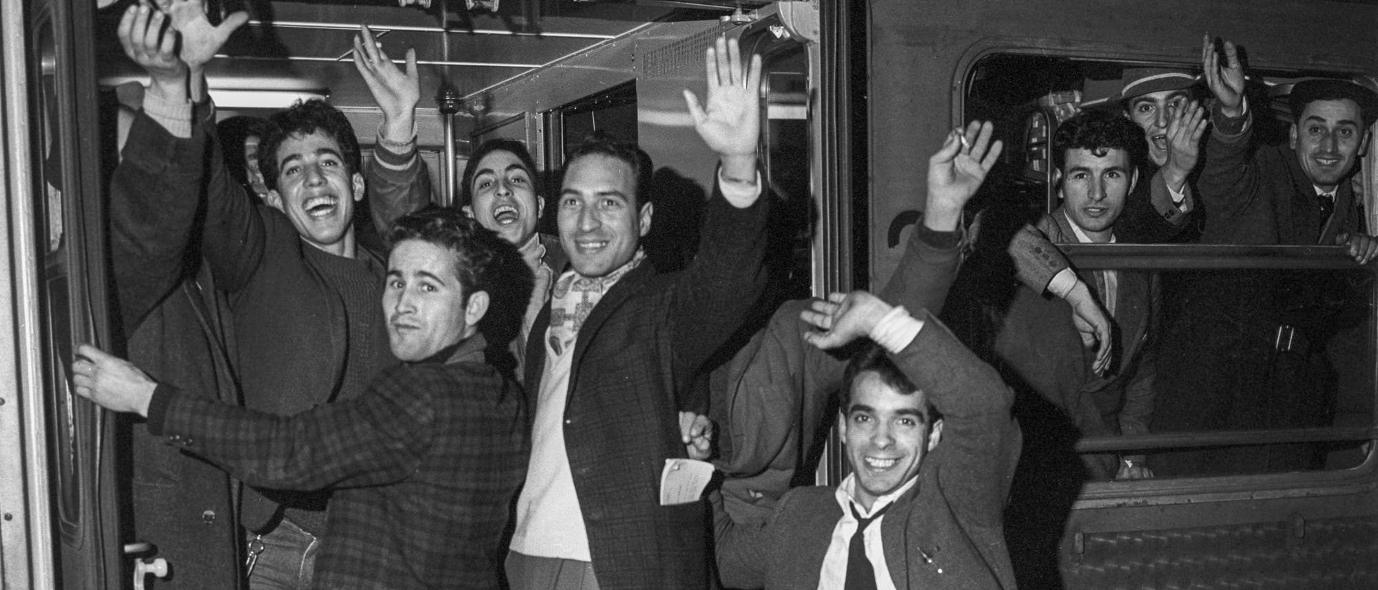 Eine Gruppe von Menschen steht in einem Zug und winkt fr&ouml;hlich in die Kamera. Einige Personen stehen in der offenen T&uuml;r, w&auml;hrend andere aus dem Fenster schauen. Die Szene wirkt lebhaft und zeigt eine positive Stimmung. Der Zug scheint an einem Bahnhof zu stehen.