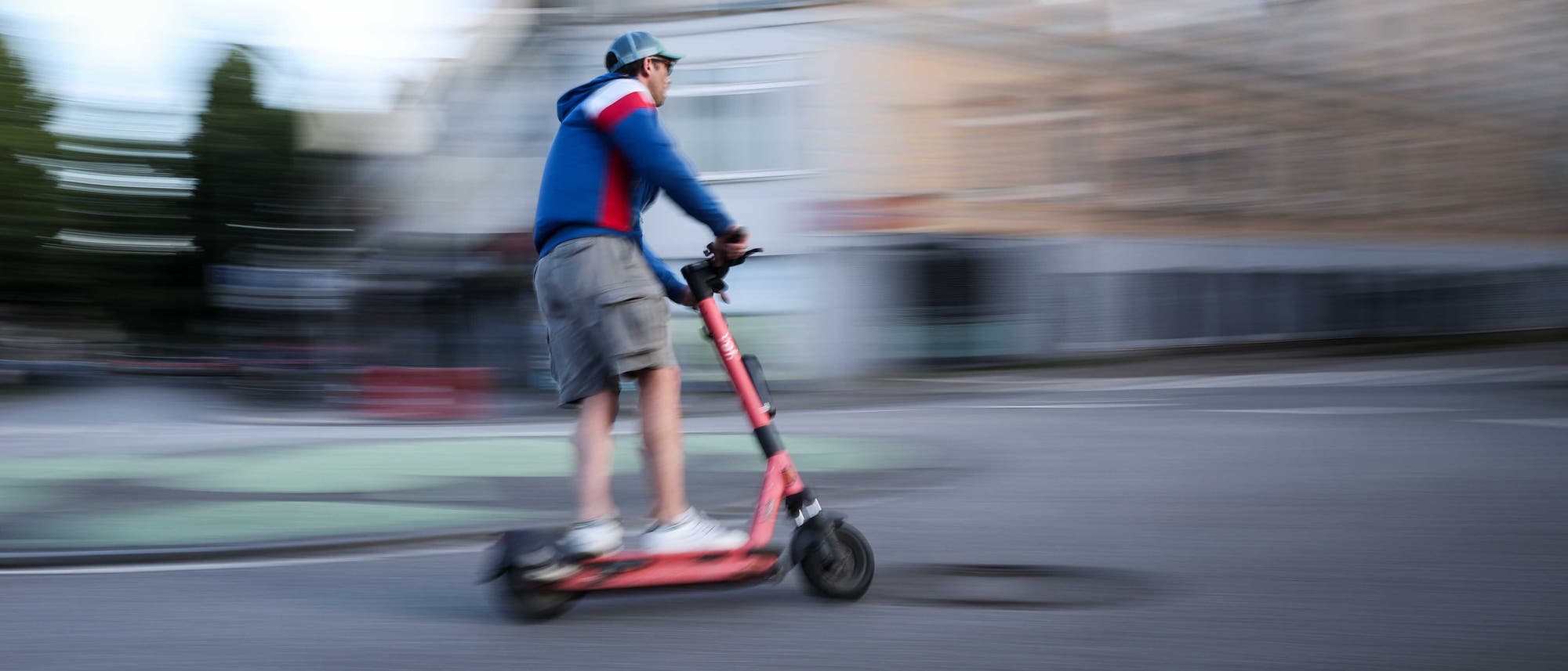 Eine Person fährt schnell auf einem roten E-Scooter durch eine städtische Umgebung. Die Bewegung ist verschwommen dargestellt, was die Geschwindigkeit betont. Die Person trägt eine blaue Kapuzenjacke, eine kurze Hose und eine Mütze. Im Hintergrund sind unscharfe Gebäude und Bäume zu sehen. Eine Person fährt schnell auf einem roten E-Scooter durch eine städtische Umgebung. Die Bewegung ist verschwommen dargestellt, was die Geschwindigkeit betont. Die Person trägt eine blaue Kapuzenjacke, eine kurze Hose und eine Mütze. Im Hintergrund sind unscharfe Gebäude und Bäume zu sehen.