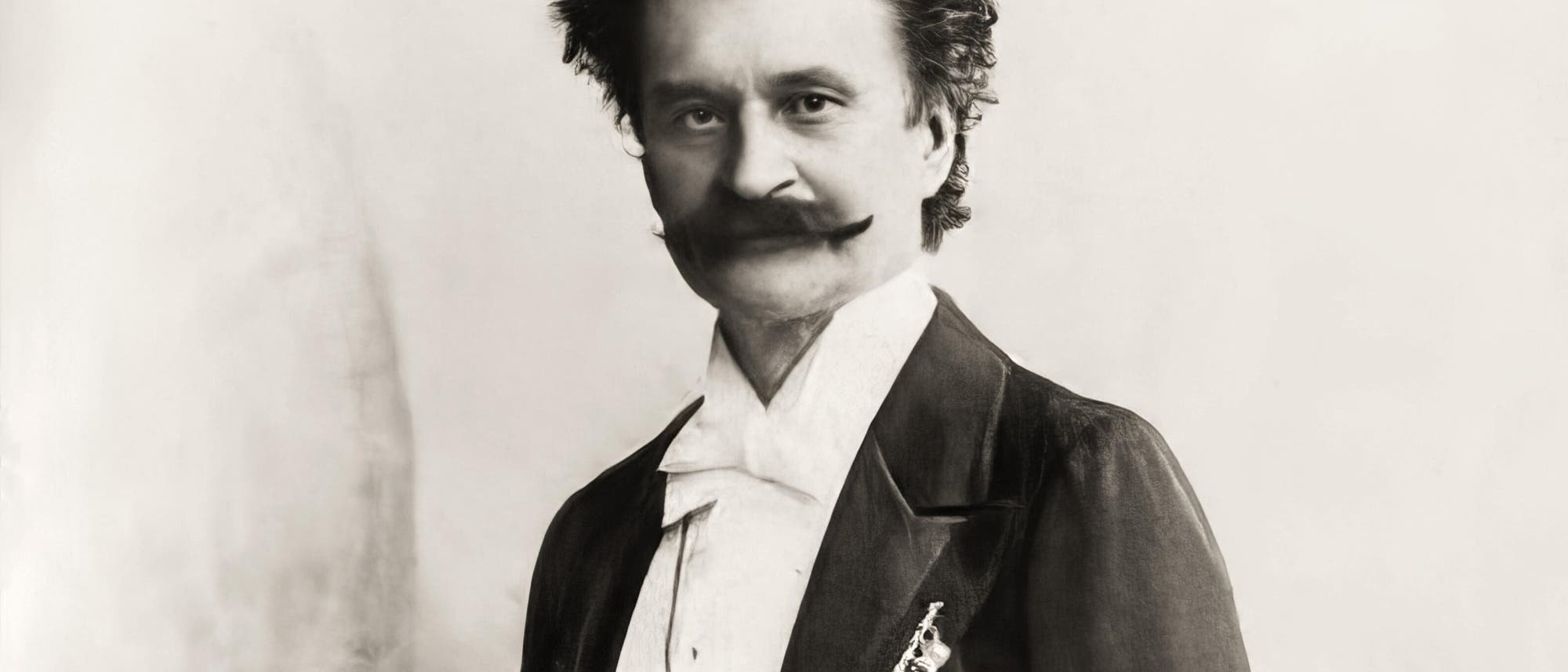 Ein schwarz-weißes Porträtfoto eines Mannes in formeller Kleidung. Er trägt einen Anzug mit Fliege und hat einen markanten Schnurrbart. Der Hintergrund ist schlicht und hell, was den Fokus auf die Person lenkt. Ein schwarz-weißes Porträtfoto eines Mannes in formeller Kleidung. Er trägt einen Anzug mit Fliege und hat einen markanten Schnurrbart. Der Hintergrund ist schlicht und hell, was den Fokus auf die Person lenkt.