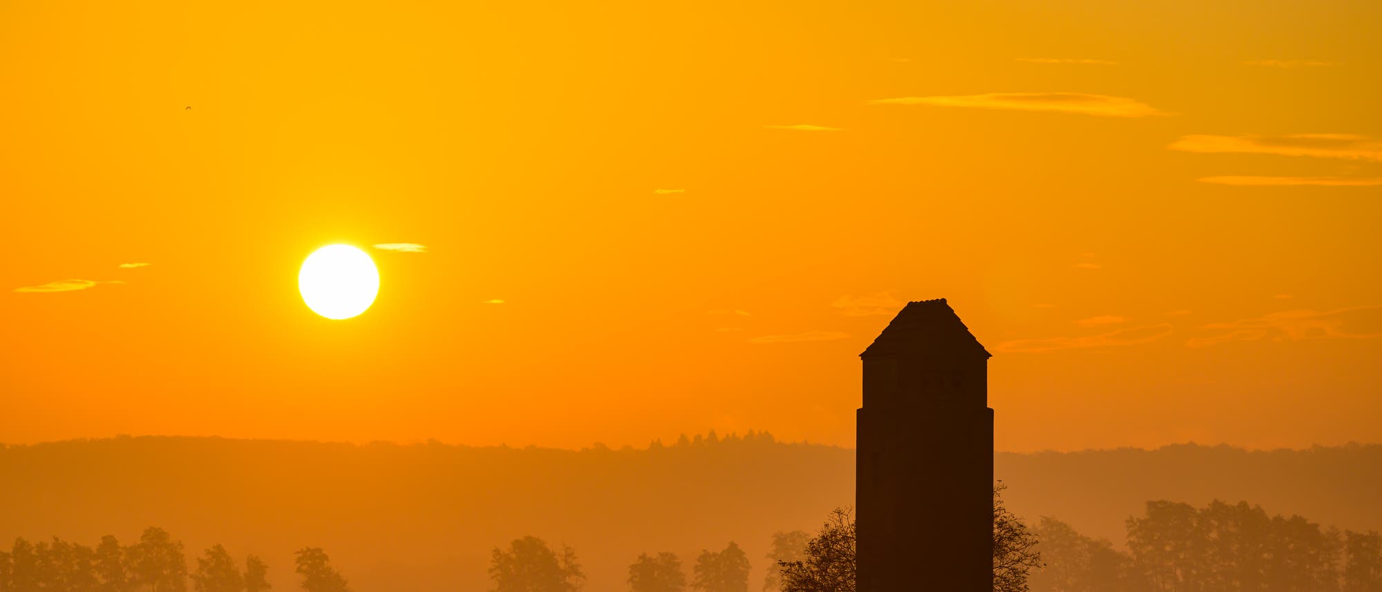 Die Sonne geht als gleißende helle Scheibe im oderbruch auf, der Himmel ist orange. Im Vordergund ist dunkel ein Turm erkennbar. Im Hintergrund befinden sich Bäume im Morgendunst, ganz hinten zeichnet sich ein Höhenzug ab