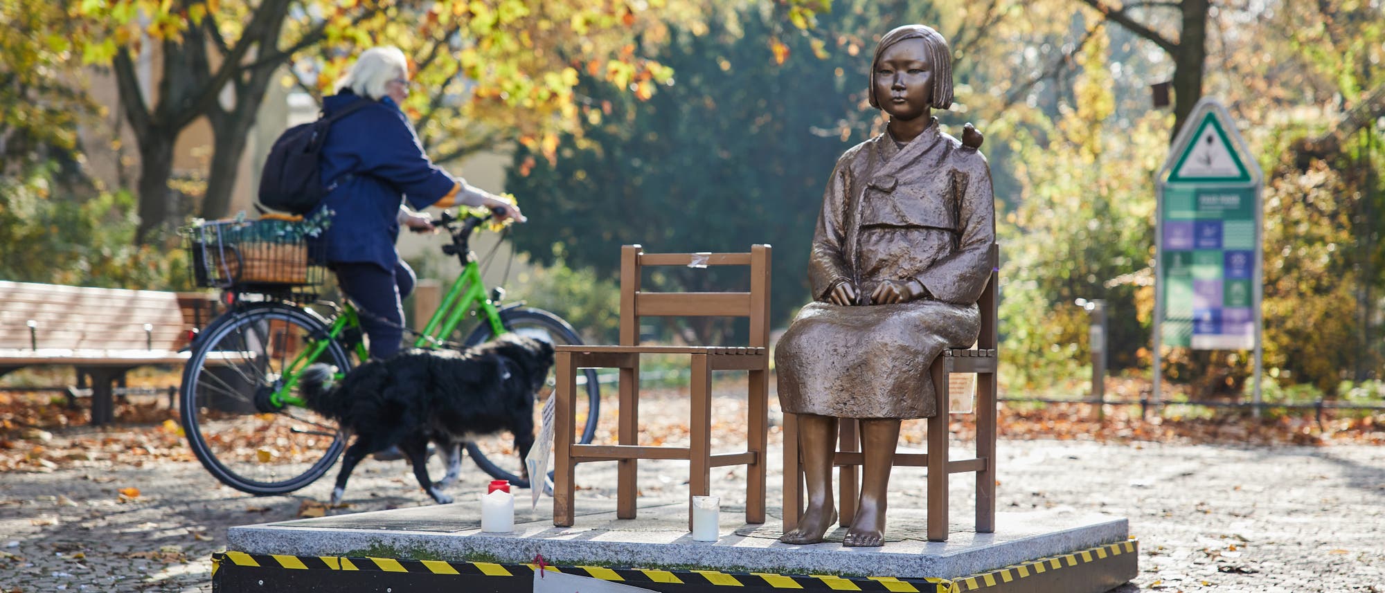 Eine Bronzestatue eines sitzenden Mädchens auf einem Podest in einem herbstlichen Park. Neben der Statue steht ein leerer Stuhl. Im Hintergrund fährt eine Person auf einem grünen Fahrrad vorbei, begleitet von einem schwarzen Hund. Auf dem Podest ist ein handgeschriebenes Schild mit der Aufschrift "ARI bleibt!" angebracht. Bäume mit herbstlichem Laub und ein Informationsschild sind im Hintergrund zu sehen.