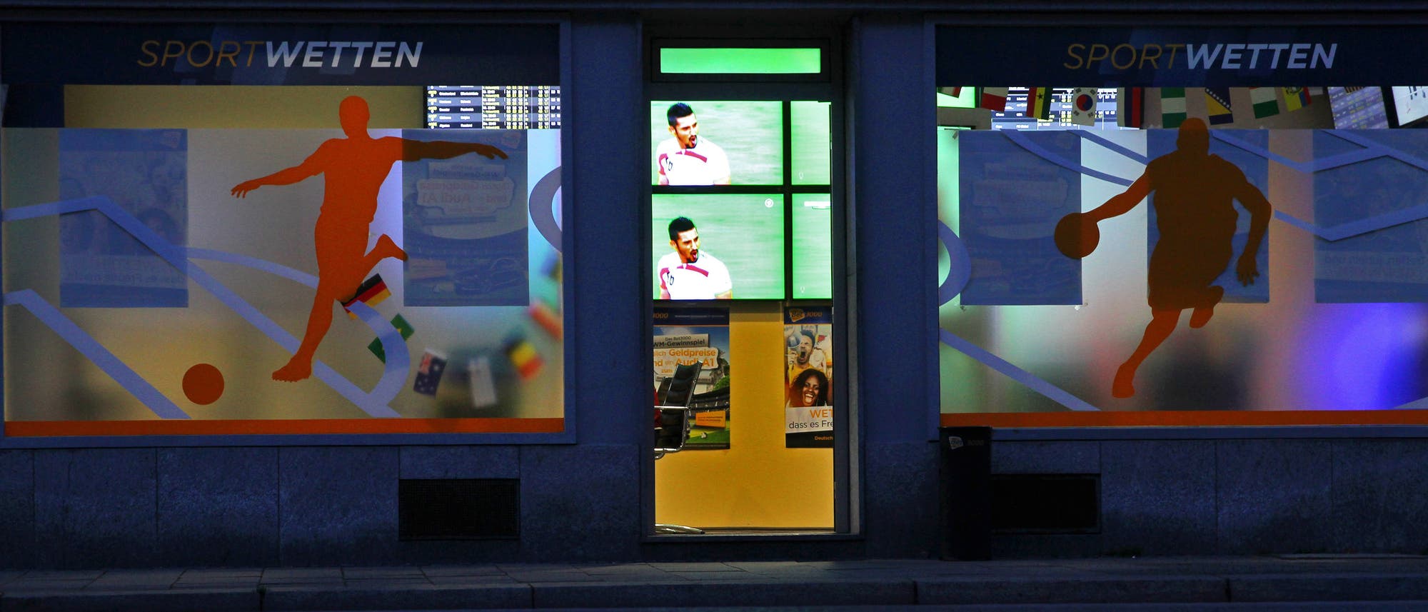 Das Foto zeigt ein erleuchtetes Wettbüro in einer nächtlichen Straße. Durch die offene oder gläserne Tür sieht man Monitore mit einer Sportübertragung.