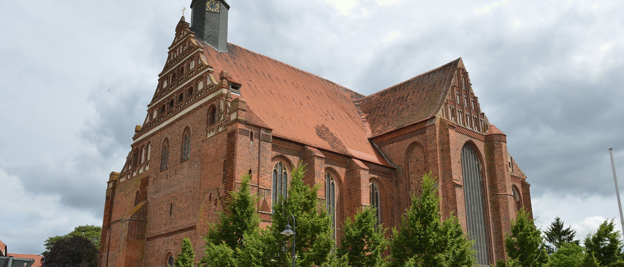 Eine gro&szlig;e Backsteinkirche mit einem hohen, spitzen Turm und einem roten Ziegeldach steht im Mittelpunkt des Bildes. Vor der Kirche sind mehrere B&auml;ume und ein kleiner Platz mit B&auml;nken und Blumenbeeten zu sehen. Der Himmel ist bew&ouml;lkt.