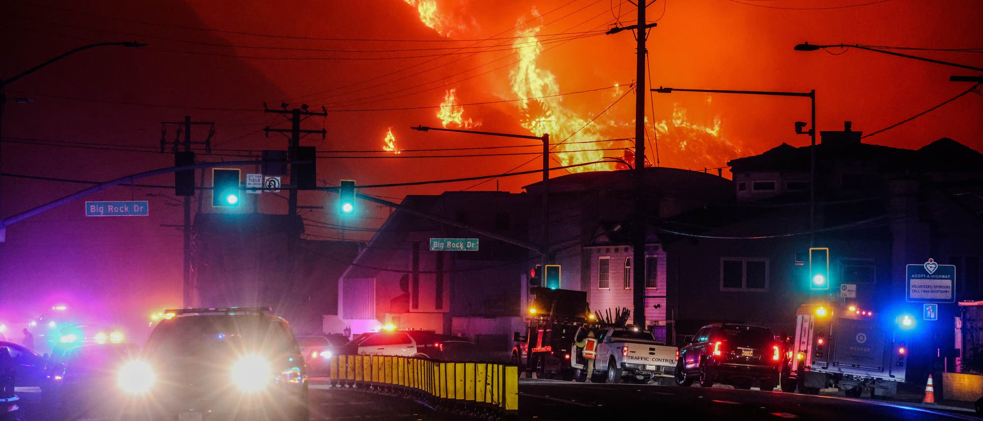 Brennender Berghang hinter Gebäuden an einer Straße mit Absperrungen und Feuerwehrfahrzeugen.