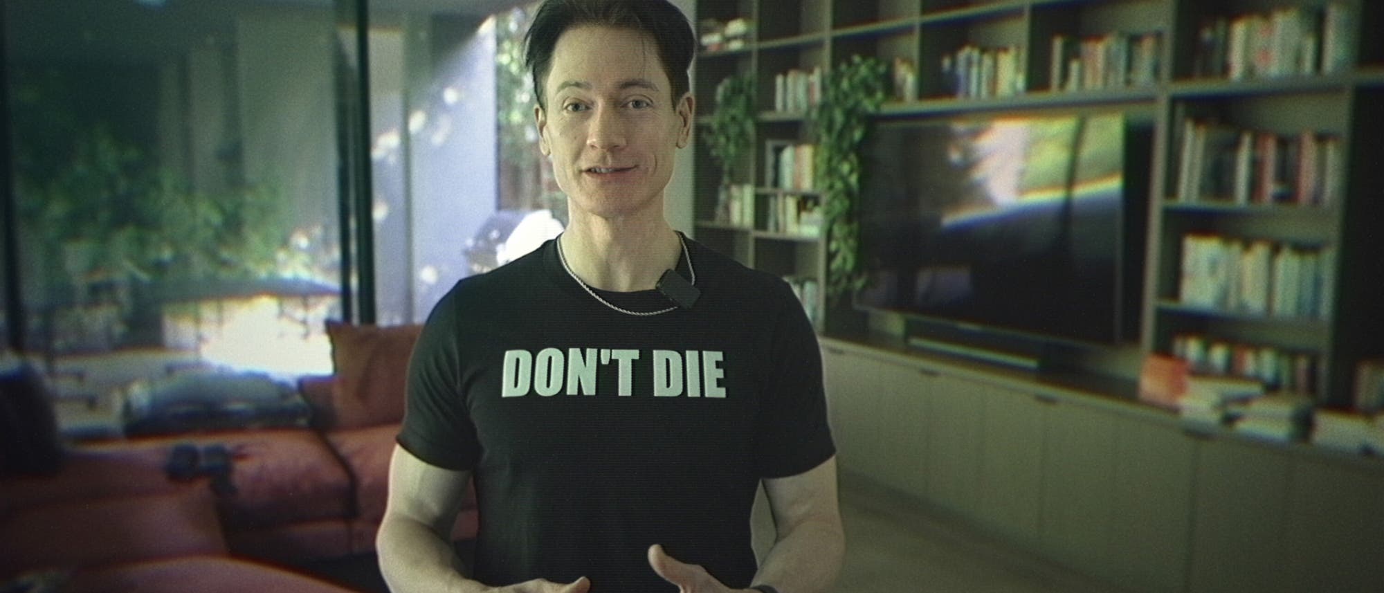 Bryan Johnson steht in einem modernen Wohnzimmer mit einem Bücherregal und einem Fernseher im Hintergrund. Er trägt ein schwarzes T-Shirt mit der Aufschrift "DON’T DIE". Bryan Johnson steht in einem modernen Wohnzimmer mit einem Bücherregal und einem Fernseher im Hintergrund. Er trägt ein schwarzes T-Shirt mit der Aufschrift "DON’T DIE".