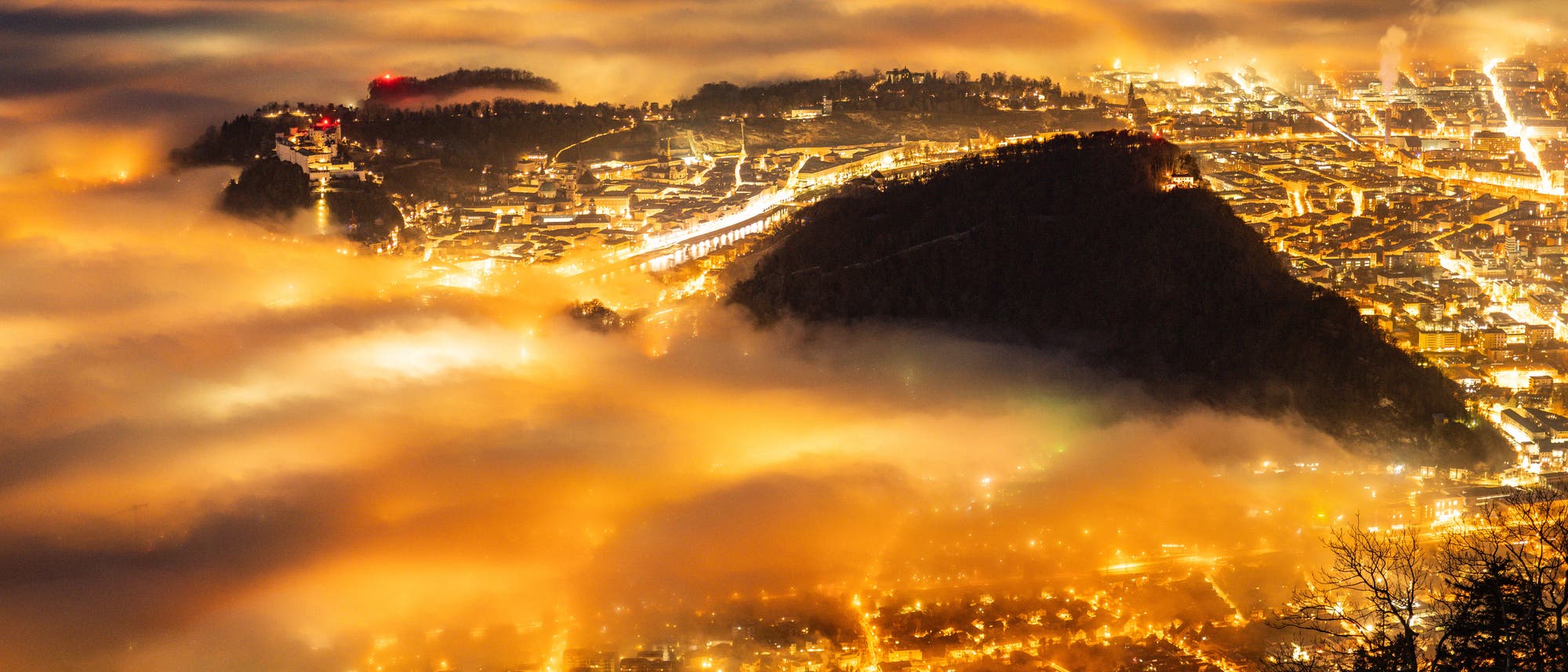 Eine nächtliche Stadtansicht, die von oben aufgenommen wurde, zeigt eine Stadt, die in warmes, goldenes Licht getaucht ist. Dichter Nebel schwebt über der Stadt und erzeugt eine mystische Atmosphäre. Die Lichter der Stadt leuchten durch den Nebel hindurch, während im Hintergrund Hügel oder Berge zu sehen sind. Die Szene vermittelt ein Gefühl von Ruhe und Erhabenheit. Eine nächtliche Stadtansicht, die von oben aufgenommen wurde, zeigt eine Stadt, die in warmes, goldenes Licht getaucht ist. Dichter Nebel schwebt über der Stadt und erzeugt eine mystische Atmosphäre. Die Lichter der Stadt leuchten durch den Nebel hindurch, während im Hintergrund Hügel oder Berge zu sehen sind. Die Szene vermittelt ein Gefühl von Ruhe und Erhabenheit.