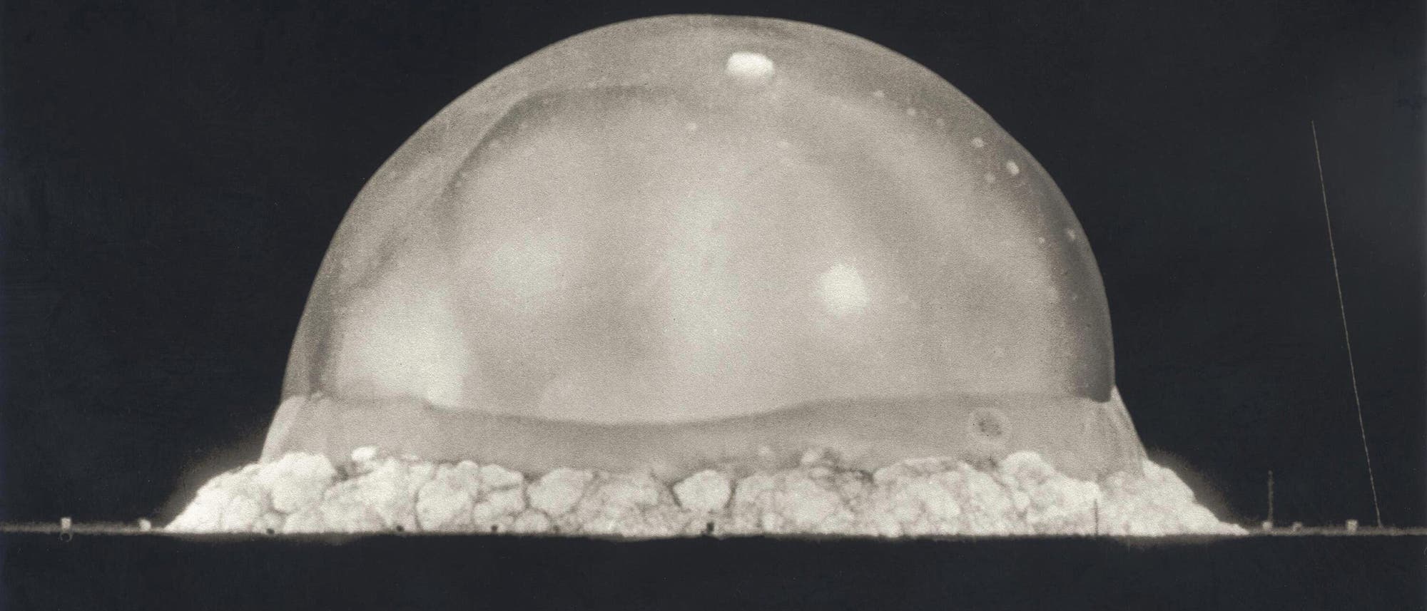 Eine Schwarz-Weiß-Fotografie zeigt die frühe Phase einer Explosion mit einer großen, halbkreisförmigen Rauchwolke, die sich über dem Boden ausbreitet. Am unteren Rand des Bildes sind die Beschriftungen „0.025 SEC“ und „100 METERS“ zu sehen, die auf die Zeit seit Beginn der Explosion und die Breite der Wolke hinweisen. Die Szene ist in einem dunklen Hintergrund eingefangen, was den Kontrast zur hellen Wolke verstärkt. Eine Schwarz-Weiß-Fotografie zeigt die frühe Phase einer Explosion mit einer großen, halbkreisförmigen Rauchwolke, die sich über dem Boden ausbreitet. Am unteren Rand des Bildes sind die Beschriftungen „0.025 SEC“ und „100 METERS“ zu sehen, die auf die Zeit seit Beginn der Explosion und die Breite der Wolke hinweisen. Die Szene ist in einem dunklen Hintergrund eingefangen, was den Kontrast zur hellen Wolke verstärkt.