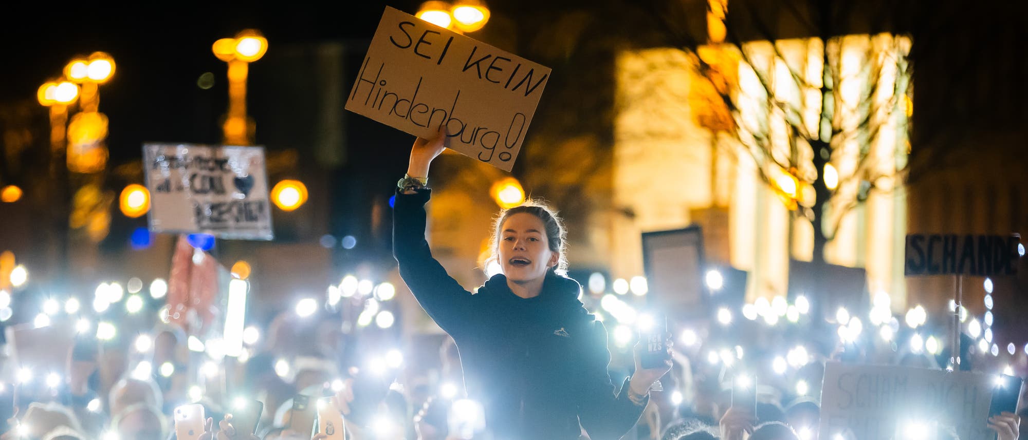 Eine junge Frau steht inmitten einer Menschenmenge bei Nacht und hält ein Schild mit der Aufschrift "Sei kein Hindenburg!" hoch. Die umstehenden Personen leuchten mit ihren Handys, wodurch ein Meer aus Licht entsteht. Im Hintergrund sind beleuchtete Straßenlaternen und unscharfe Gebäude zu sehen. Die Szene vermittelt den Eindruck einer friedlichen, aber emotional aufgeladenen Demonstration oder eines Protests. Eine junge Frau steht inmitten einer Menschenmenge bei Nacht und hält ein Schild mit der Aufschrift "Sei kein Hindenburg!" hoch. Die umstehenden Personen leuchten mit ihren Handys, wodurch ein Meer aus Licht entsteht. Im Hintergrund sind beleuchtete Straßenlaternen und unscharfe Gebäude zu sehen. Die Szene vermittelt den Eindruck einer friedlichen, aber emotional aufgeladenen Demonstration oder eines Protests.