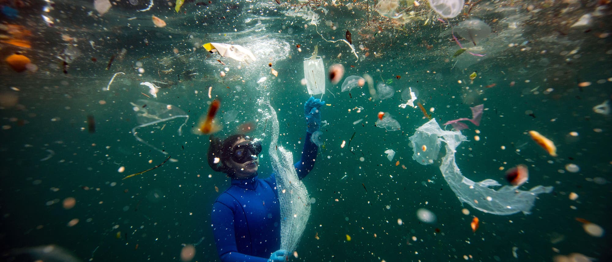 Eine Person in einem blauen Taucheranzug und einer Tauchmaske schwimmt unter Wasser inmitten von Plastikmüll. Sie hält ein Stück Plastik in der Hand, umgeben von verschiedenen Abfällen wie Plastiktüten und Flaschen. Das Bild thematisiert die Verschmutzung der Ozeane durch Plastikmüll.
