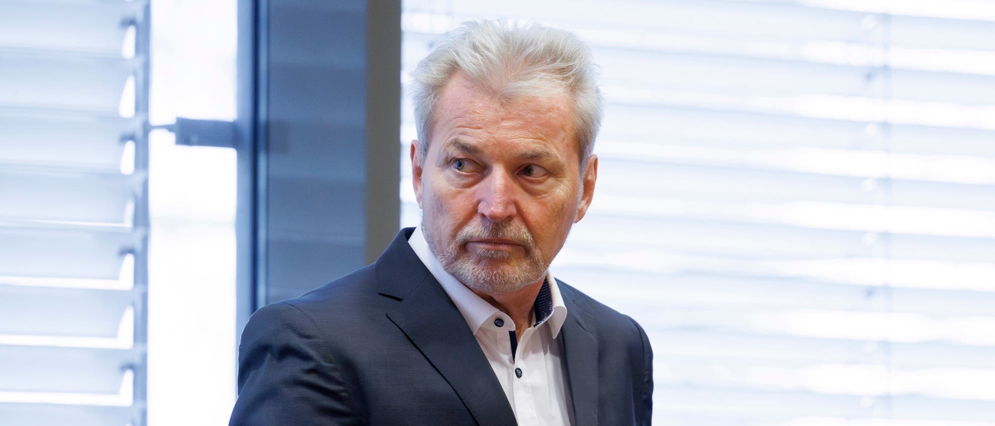 Ein Mann in einem Anzug steht in einem Raum mit gro&szlig;en Fenstern und Jalousien. Er schaut zur Seite und wirkt nachdenklich. Die Beleuchtung ist hell und der Hintergrund zeigt verschwommene Details der Fensterstruktur.