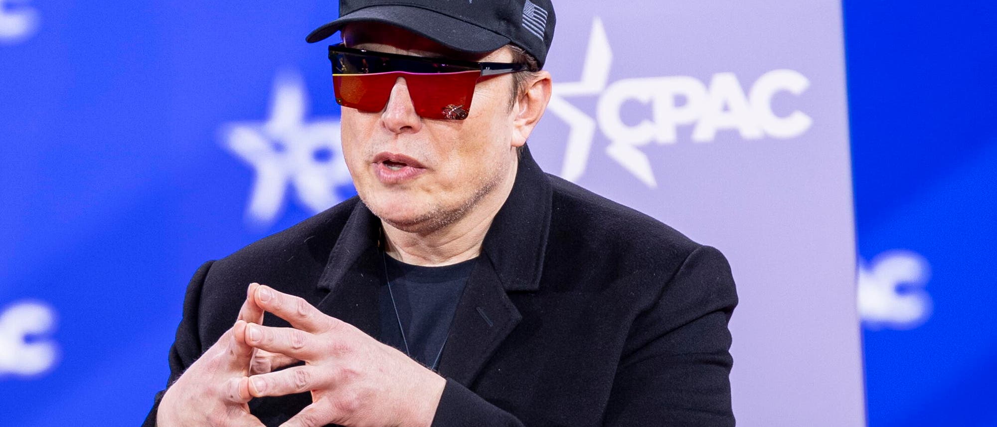 Das Gesicht eines mittfünfzigjährigen Mannes mit leichten Bartstoppeln und knubbeliger, heller Hautstruktur wird von einer futuristischen Sonnenbrille, einer schwarzen Schirmmütze mit US-Flagge, einem schwarzen Samtsakko und zusammengelegten Fingern umrahmt, deren Geste an diejenige von Montgomery Burns aus den Simpsons erinnert. Das Gesicht eines mittfünfzigjährigen Mannes mit leichten Bartstoppeln und knubbeliger, heller Hautstruktur wird von einer futuristischen Sonnenbrille, einer schwarzen Schirmmütze mit US-Flagge, einem schwarzen Samtsakko und zusammengelegten Fingern umrahmt, deren Geste an diejenige von Montgomery Burns aus den Simpsons erinnert.