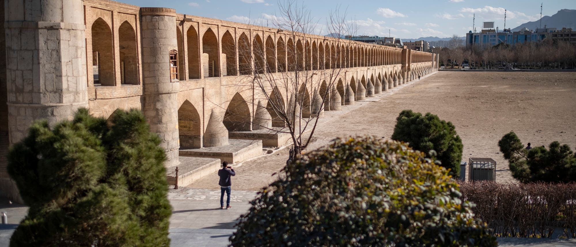 Eine historische Brücke mit mehreren Bögen erstreckt sich über ein trockenes Flussbett. Die Brücke besteht aus hellem Stein und zeigt eine Reihe von Arkaden. Im Vordergrund sind Bäume und ein Mensch, der die Brücke fotografiert. Der Himmel ist klar mit einigen Wolken, und im Hintergrund sind Gebäude und Berge zu sehen. Eine historische Brücke mit mehreren Bögen erstreckt sich über ein trockenes Flussbett. Die Brücke besteht aus hellem Stein und zeigt eine Reihe von Arkaden. Im Vordergrund sind Bäume und ein Mensch, der die Brücke fotografiert. Der Himmel ist klar mit einigen Wolken, und im Hintergrund sind Gebäude und Berge zu sehen.