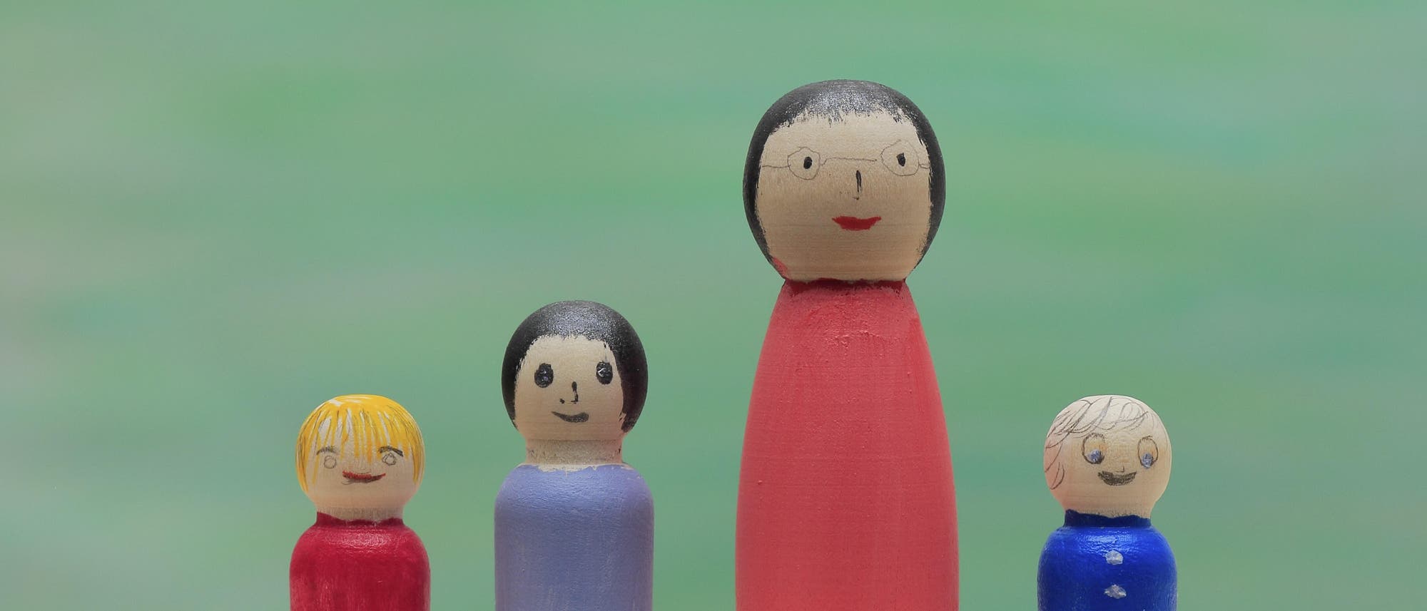 Eine Gruppe von vier bemalten Holzfiguren steht auf einer Holzoberfl&auml;che vor einem verschwommenen gr&uuml;nen Hintergrund. Die Figuren variieren in Gr&ouml;&szlig;e und Farbe: eine gro&szlig;e Figur in Rot, zwei kleinere in Blau und eine kleinere ebenfalls in Rot. Jede Figur hat ein einfaches, aufgemaltes Gesicht mit unterschiedlichen Haarfarben. 