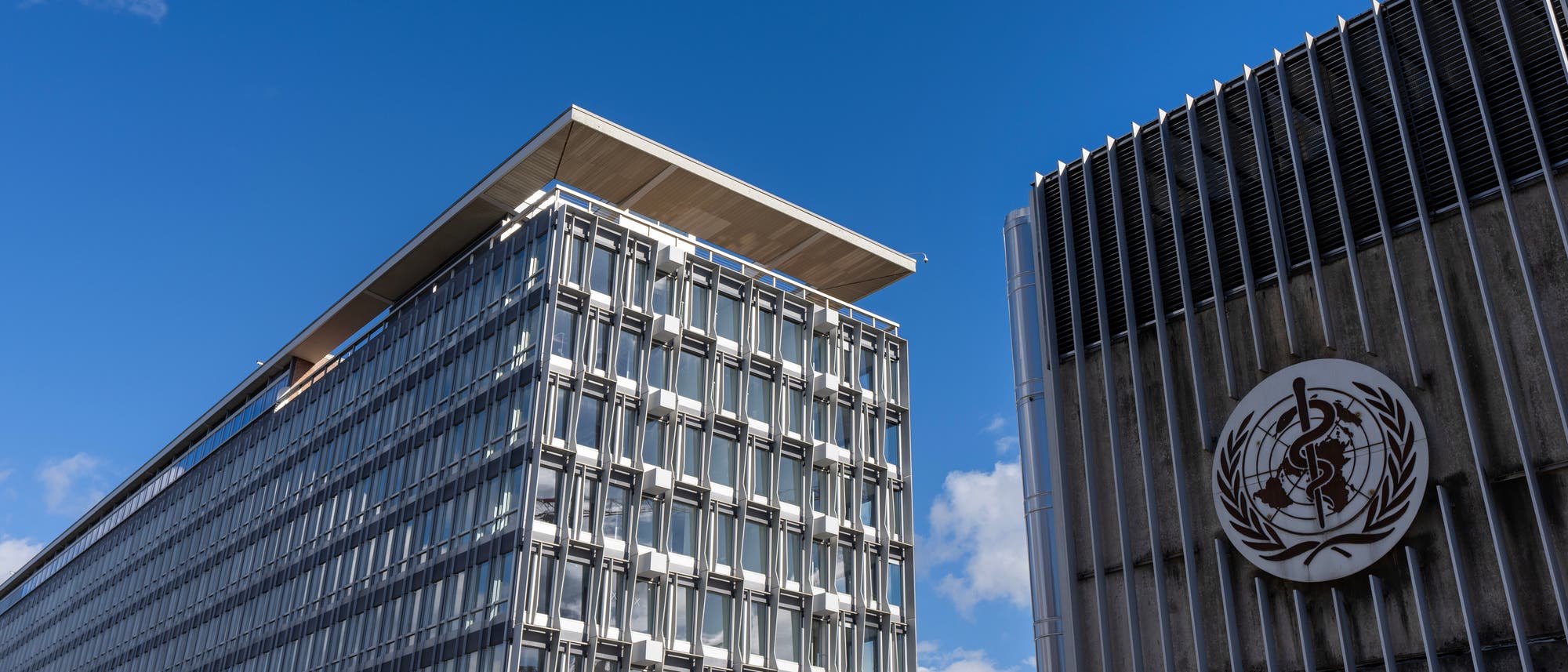 Zwei moderne Bürogebäude unter einem klaren blauen Himmel. Links steht ein hohes Gebäude mit vielen Fenstern und einer markanten Dachkante, rechts ein Gebäude mit dem Logo der Weltgesundheitsorganisation (WHO) auf der Fassade. Vor dem linken Gebäude sind mehrere internationale Flaggen zu sehen.