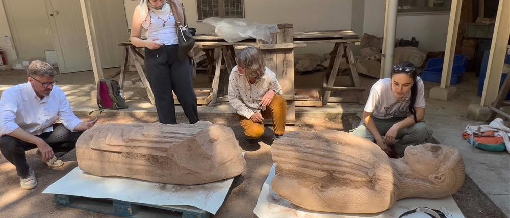 Eine Gruppe von vier Personen betrachtet zwei große, liegende Steinskulpturen, die auf Holzpaletten im Freien platziert sind. Die Skulpturen scheinen historische oder kulturelle Artefakte zu sein. Die Personen stehen oder hocken um die Skulpturen herum und scheinen in eine Diskussion vertieft zu sein. Im Hintergrund sind Werkbänke und Werkzeuge zu sehen, was auf eine archäologische oder restaurative Umgebung hindeutet. Eine Gruppe von vier Personen betrachtet zwei große, liegende Steinskulpturen, die auf Holzpaletten im Freien platziert sind. Die Skulpturen scheinen historische oder kulturelle Artefakte zu sein. Die Personen stehen oder hocken um die Skulpturen herum und scheinen in eine Diskussion vertieft zu sein. Im Hintergrund sind Werkbänke und Werkzeuge zu sehen, was auf eine archäologische oder restaurative Umgebung hindeutet.