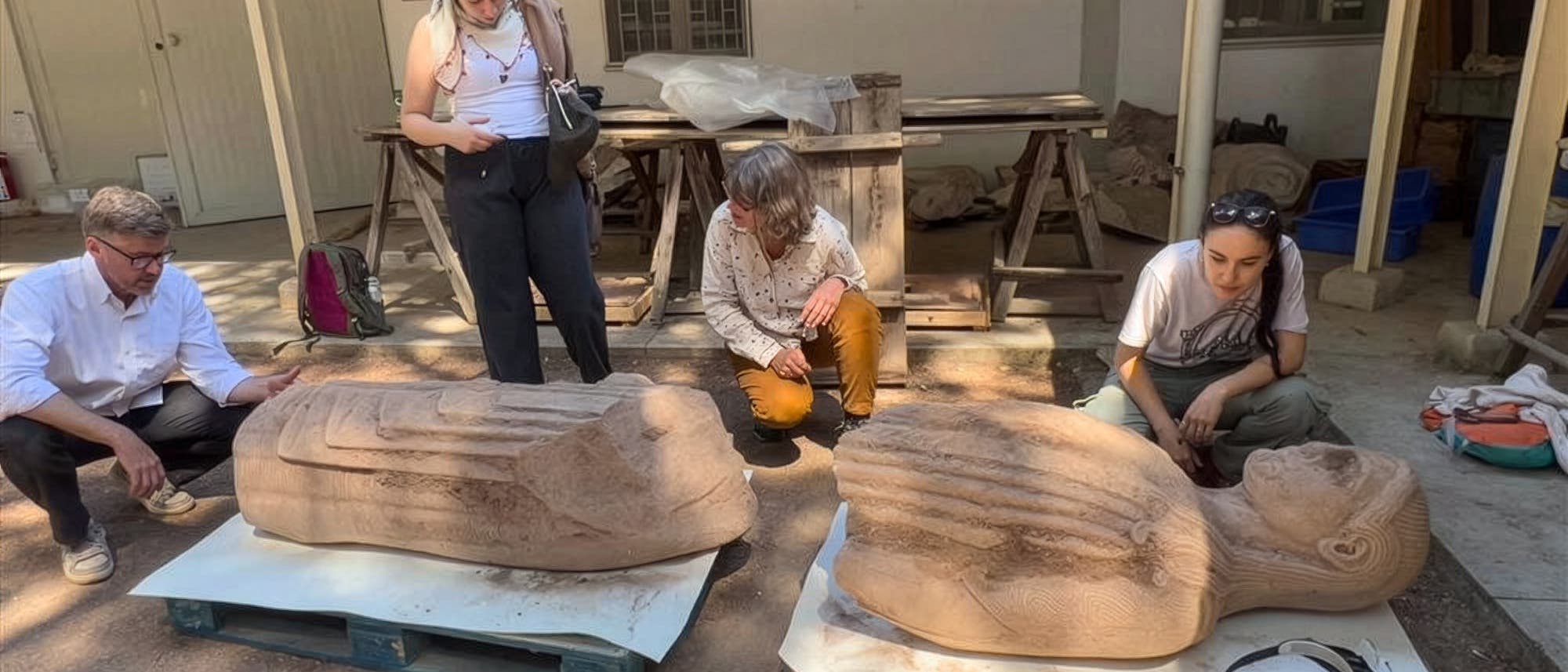 Eine Gruppe von vier Personen betrachtet zwei große, liegende Steinskulpturen, die auf Holzpaletten im Freien platziert sind. Die Skulpturen scheinen historische oder kulturelle Artefakte zu sein. Die Personen stehen oder hocken um die Skulpturen herum und scheinen in eine Diskussion vertieft zu sein. Im Hintergrund sind Werkbänke und Werkzeuge zu sehen, was auf eine archäologische oder restaurative Umgebung hindeutet. Eine Gruppe von vier Personen betrachtet zwei große, liegende Steinskulpturen, die auf Holzpaletten im Freien platziert sind. Die Skulpturen scheinen historische oder kulturelle Artefakte zu sein. Die Personen stehen oder hocken um die Skulpturen herum und scheinen in eine Diskussion vertieft zu sein. Im Hintergrund sind Werkbänke und Werkzeuge zu sehen, was auf eine archäologische oder restaurative Umgebung hindeutet.