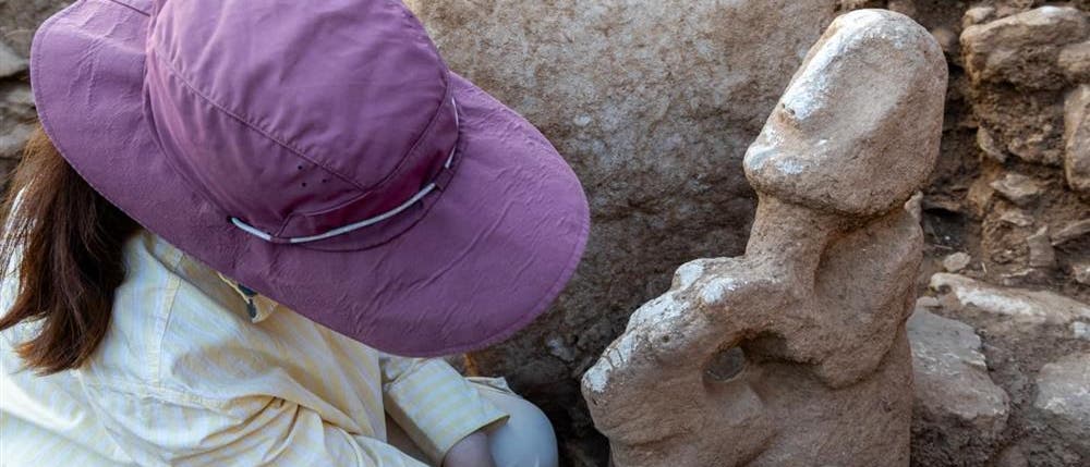 Eine Person mit einem lila Hut und einem gestreiften Hemd reinigt mit einem Pinsel eine archäologische Steinfigur. Die Szene findet in einer Ausgrabungsstätte statt, umgeben von Erde und Steinen. Die Figur hat eine abstrakte Form und scheint Teil einer größeren archäologischen Entdeckung zu sein. Eine Person mit einem lila Hut und einem gestreiften Hemd reinigt mit einem Pinsel eine archäologische Steinfigur. Die Szene findet in einer Ausgrabungsstätte statt, umgeben von Erde und Steinen. Die Figur hat eine abstrakte Form und scheint Teil einer größeren archäologischen Entdeckung zu sein.