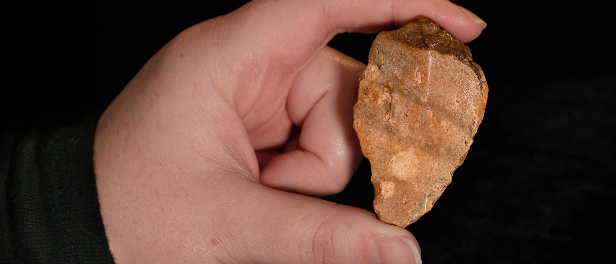 Eine Hand h&auml;lt ein steinernes Artefakt, das wie eine Pfeilspitze geformt ist, vor einem schwarzen Hintergrund. Auf der Oberfl&auml;che des Artefakts sind nat&uuml;rliche Texturen und Farbvariationen zu erkennen. 