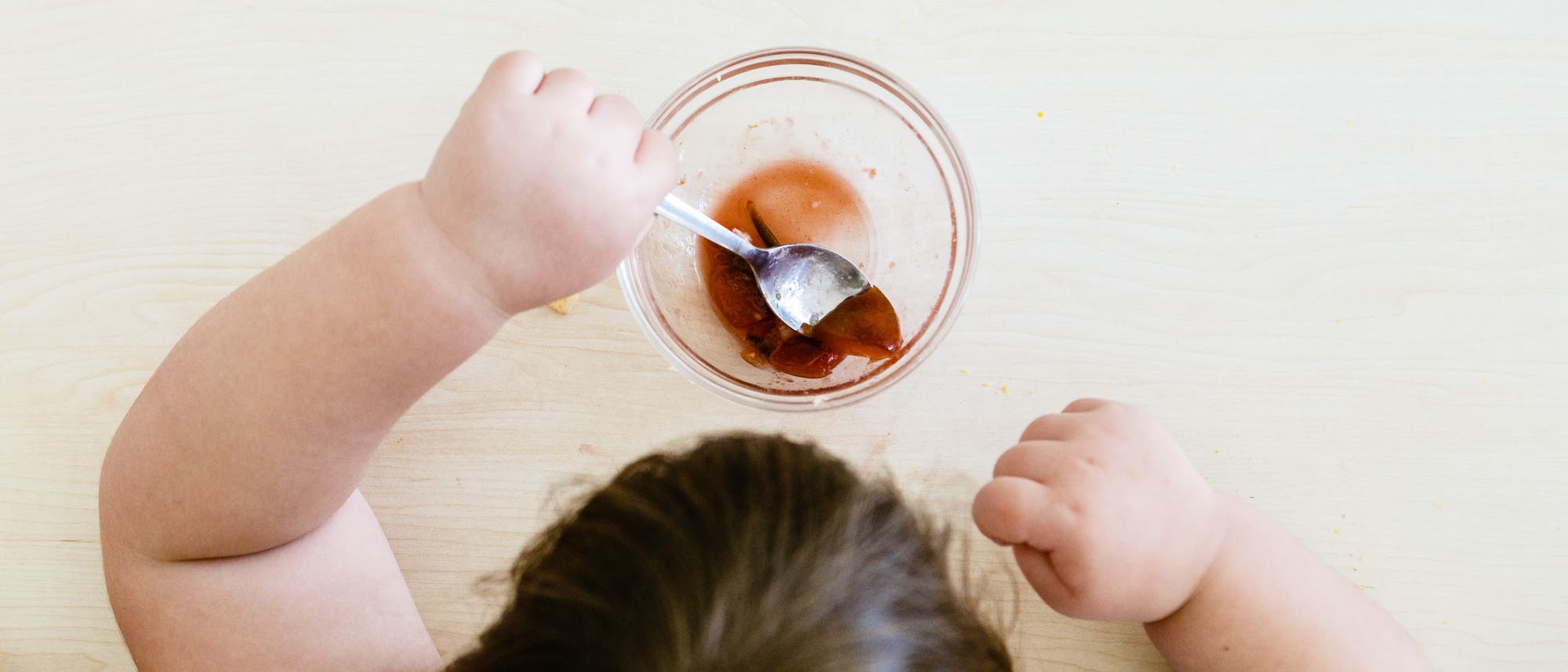 Blick von oben auf den Kopf eines Kindes mit braunen Haaren, das an an einem Holztisch sitzt und mit einem Löffel aus einer kleinen Glasschüssel eine bräunliche, an Götterspeise erinnernde Nachspeise isst. Auf dem Tisch im oberen linken Eck steht ein Glas mit einem gelben Getränk.