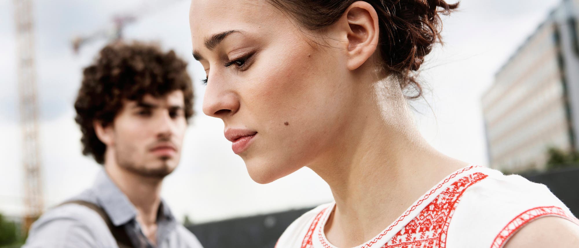 Eine junge Frau im Vordergrund blickt bedrückt nach unten, während ein Mann im Hintergrund, der sie anschaut, nur unscharf zu sehen ist. Die Frau trägt ein weißes Oberteil mit rotem Muster. Im Hintergrund sind Baukräne und ein modernes Gebäude sichtbar, was auf eine städtische Umgebung hindeutet. Die Szene vermittelt eine nachdenkliche bis krisenhafte Stimmung.